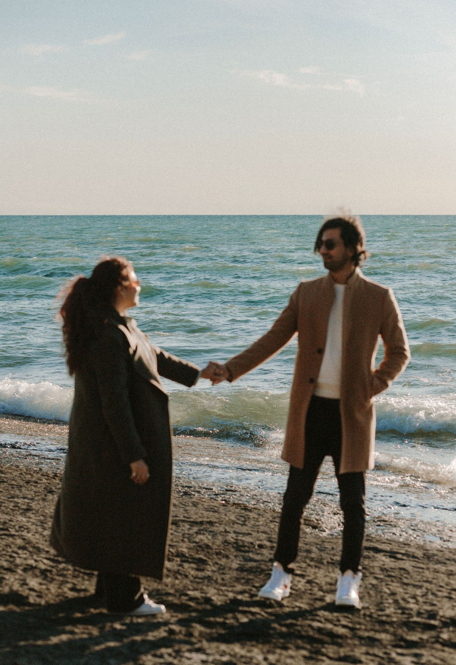A couple holds hands while walking along a scenic beach shore during a romantic autumn engagement session.
