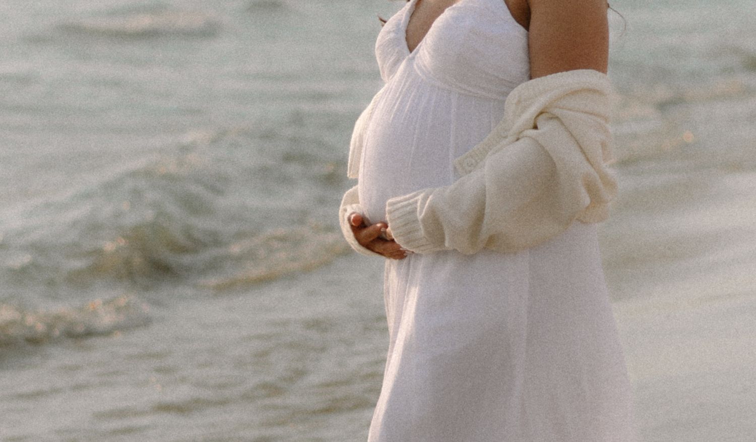 A person in a white sweater stands near the ocean waves on a cloudy day at the beach.