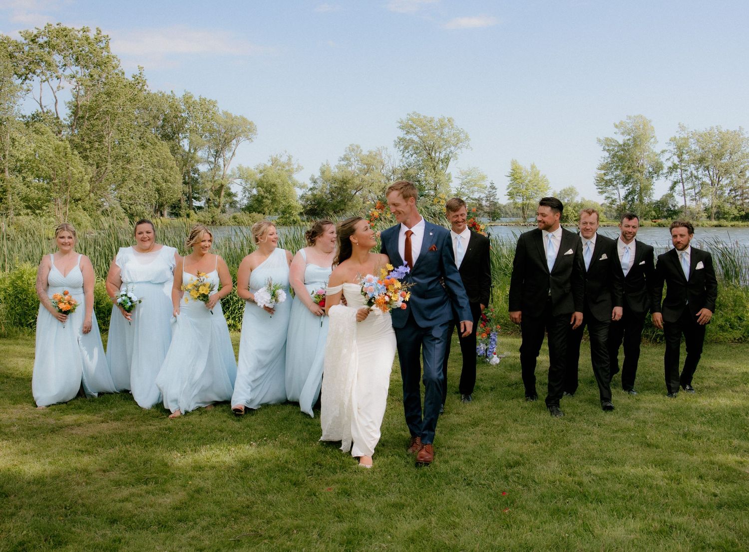 Wedding party walks outdoors near a lake, bridesmaids in light blue gowns, groomsmen in black suits, bride and groom centered.