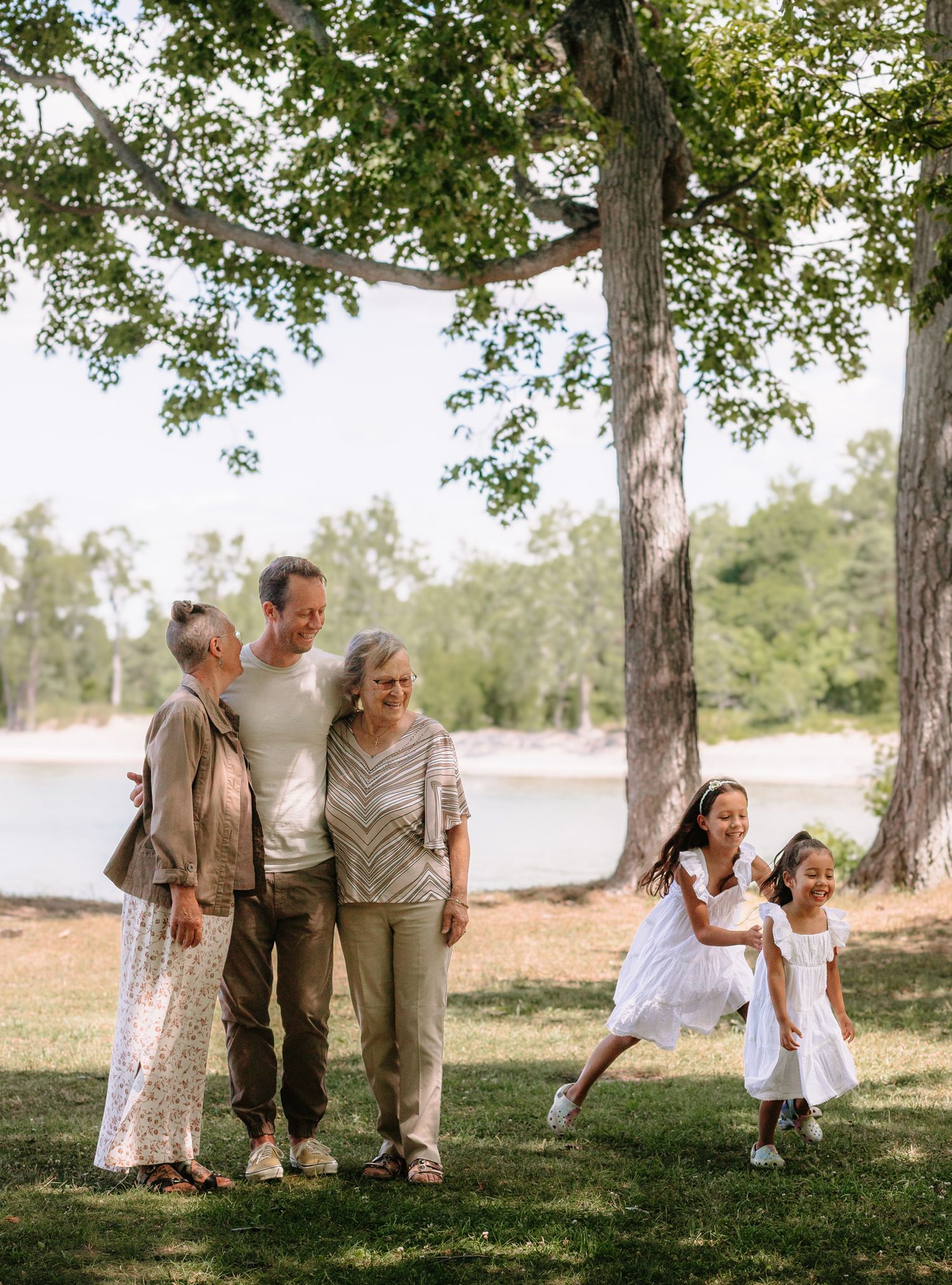A multi-generational family enjoys a sunny afternoon together by a lake under tall trees in Prince Edward County