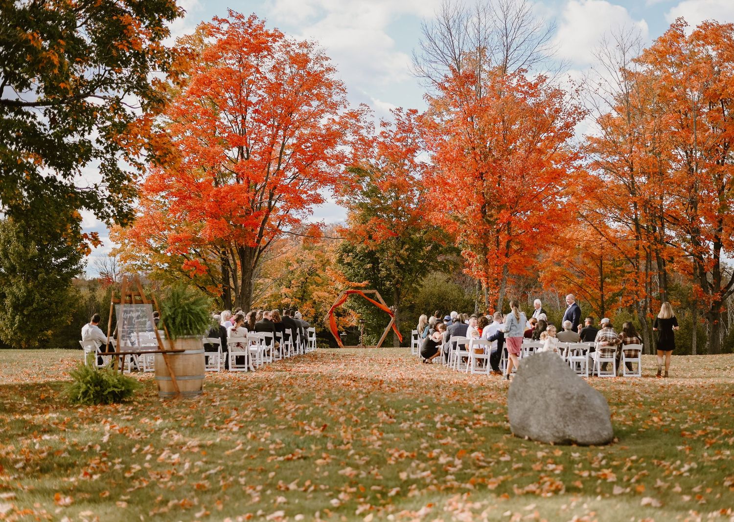 Guests gather for an intimate outdoor fall wedding ceremony surrounded by vibrant orange and red autumn foliage.