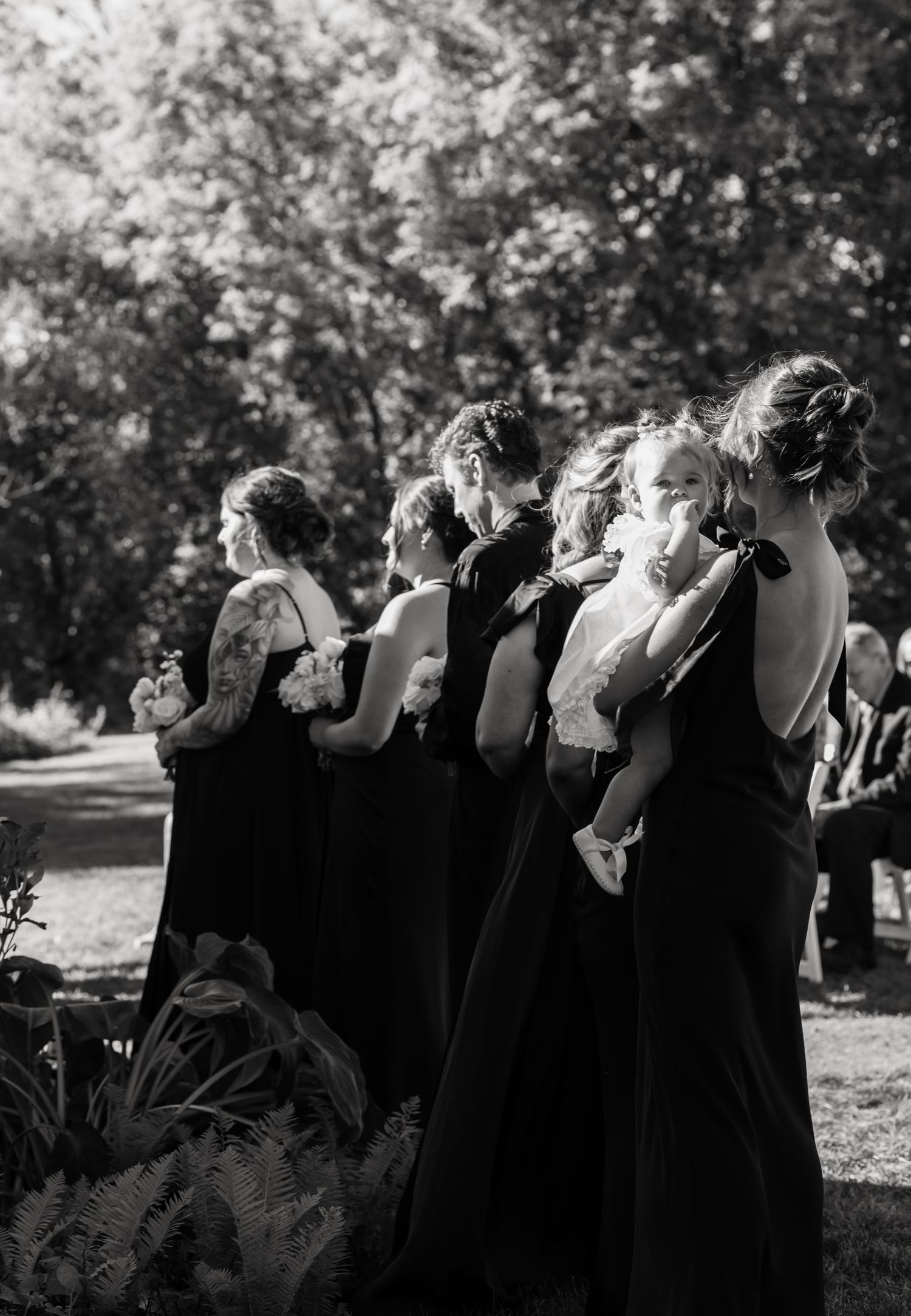 Black and white photo of bridesmaids in dark gowns watching outdoor ceremony.