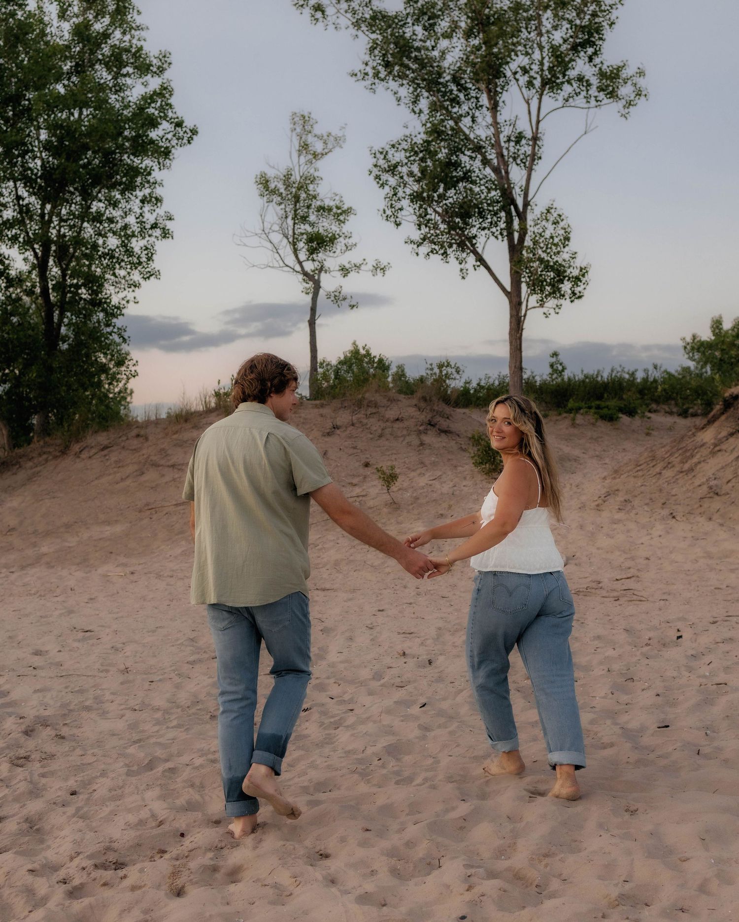 Couple holding hands and dancing on sandy beach dunes at sunset surrounded by green trees.