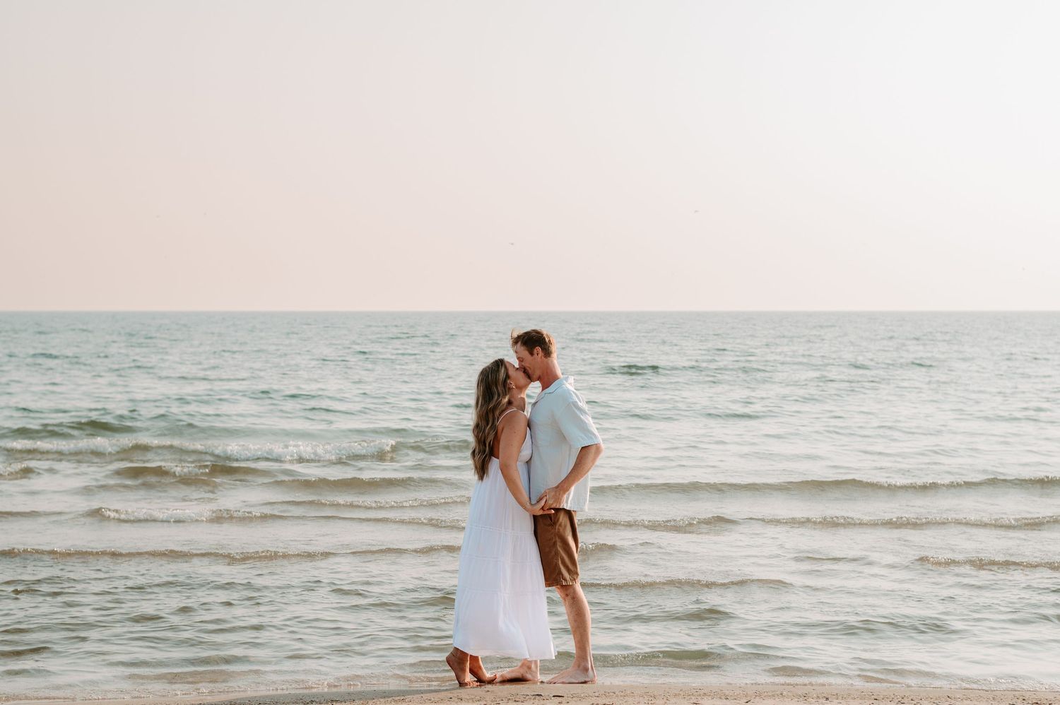 A couple shares a romantic moment on a sandy beach at sunset with waves gently rolling in the background.
