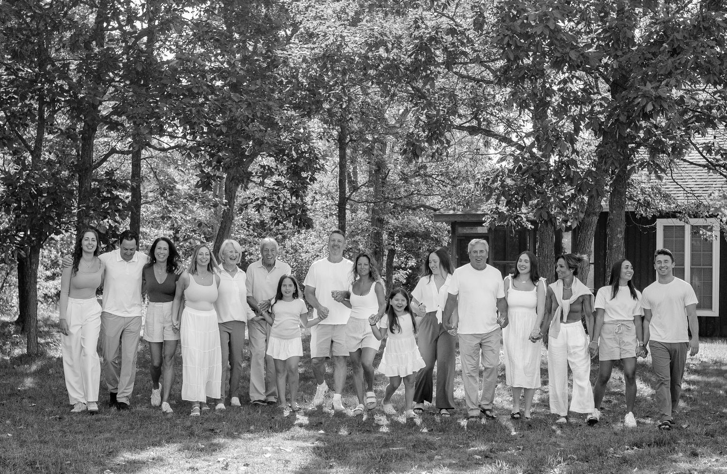 Large family group portrait outdoors in black and white, posing together among trees near a cabin.