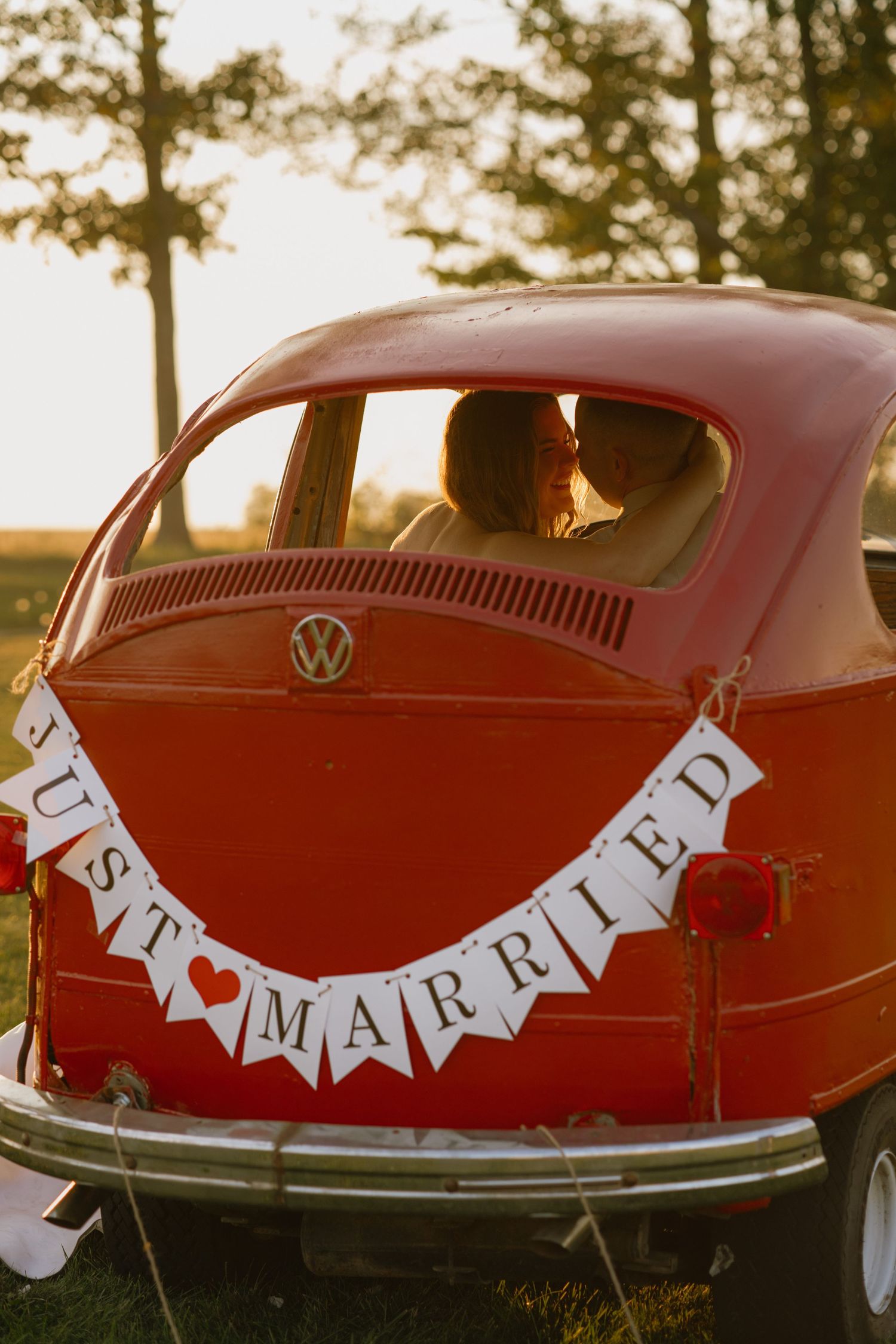 Newlyweds kiss inside a vintage red VW Beetle decorated with a Just Married banner at sunset.