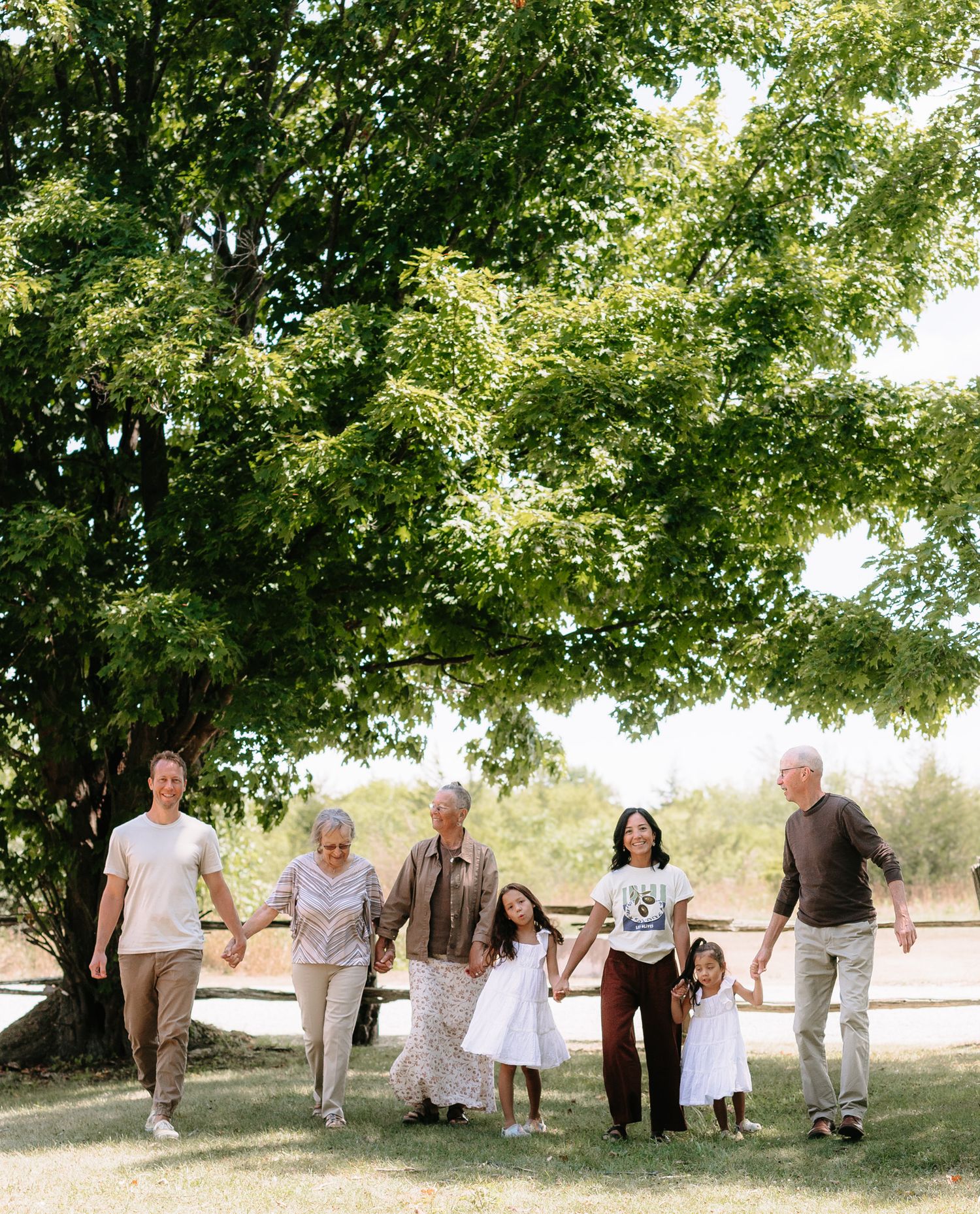 A multi-generational family walks together under a large shady tree on a sunny summer day.