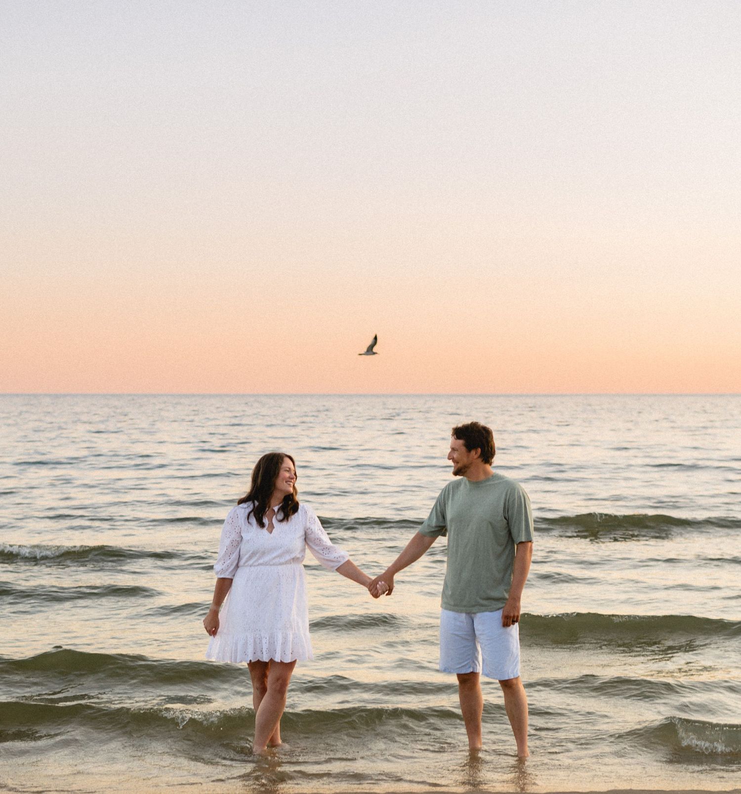A couple holds hands while wading in the ocean waves at sunset, creating a romantic silhouette against the pink sky.