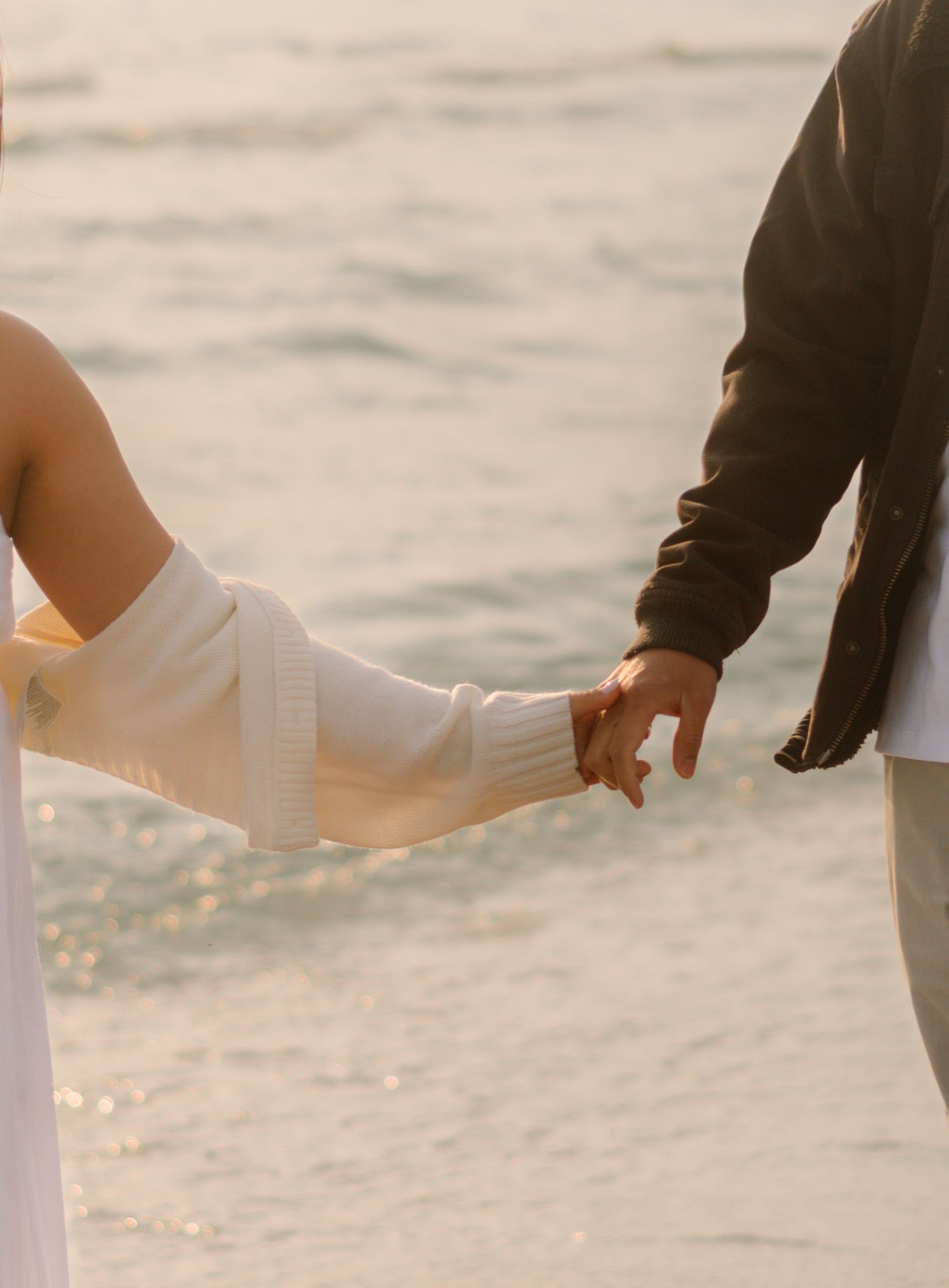 Couple holding hands while standing on a sandy beach at sunset with ocean waves in the background in Prince Edward County