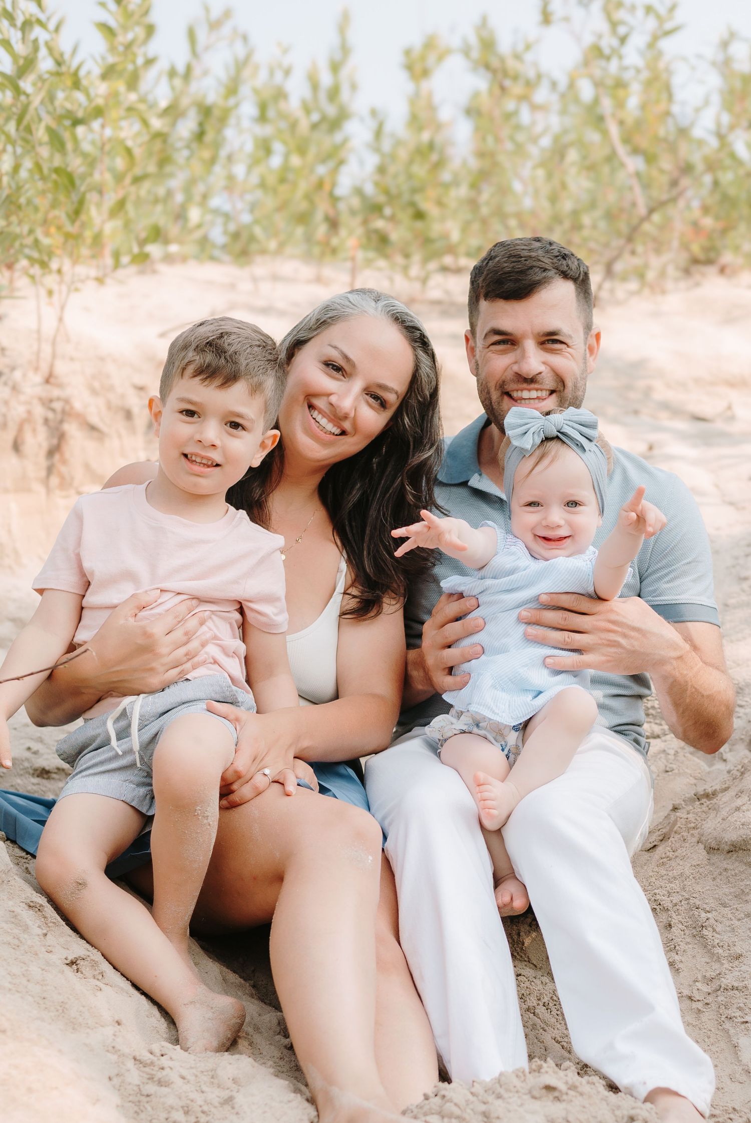 Happy family of four sitting on sandy beach, parents smiling with young boy and baby girl in blue bow.