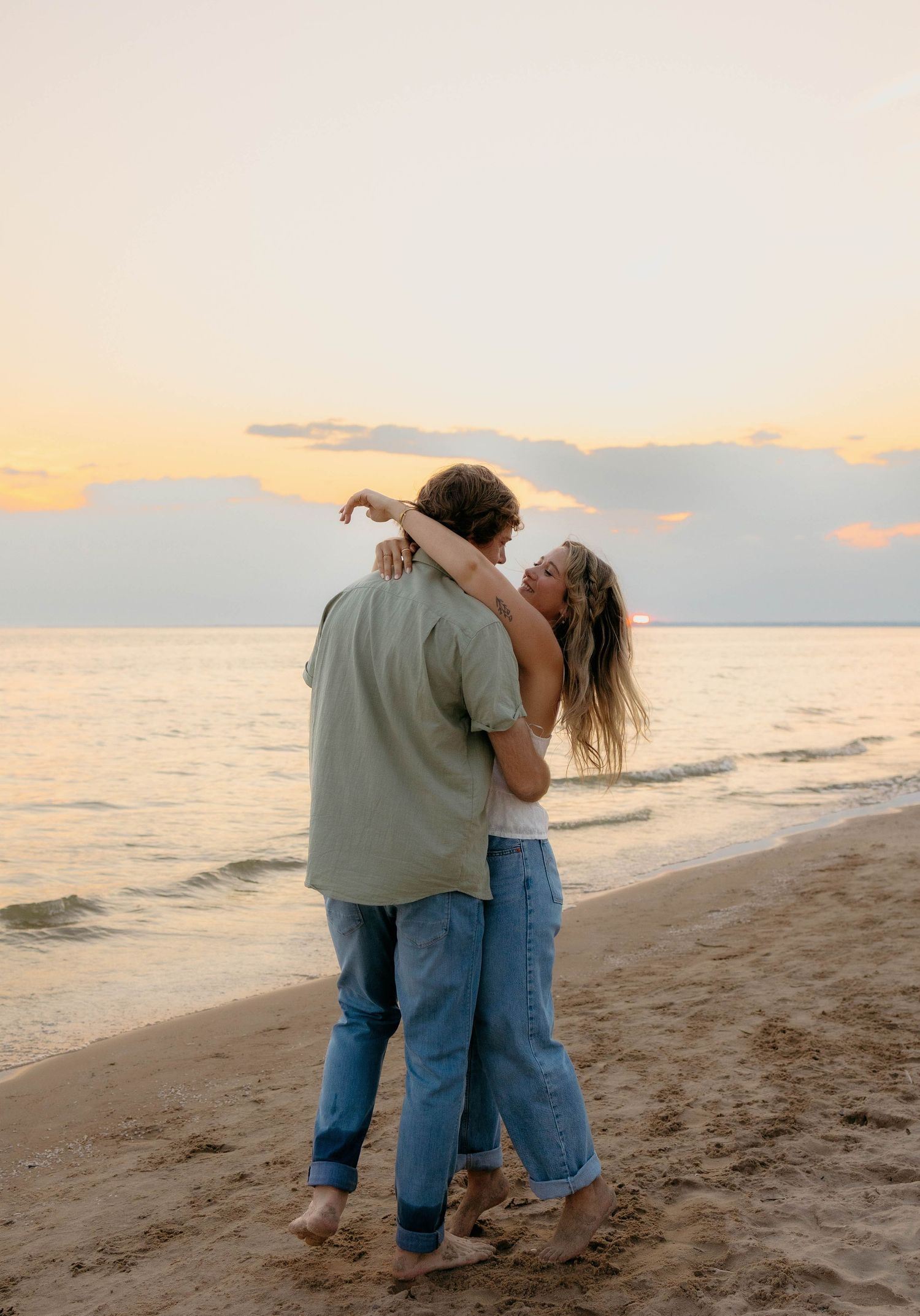 Couple dancing barefoot on a sandy beach at sunset, embracing romantically with ocean waves in the background.