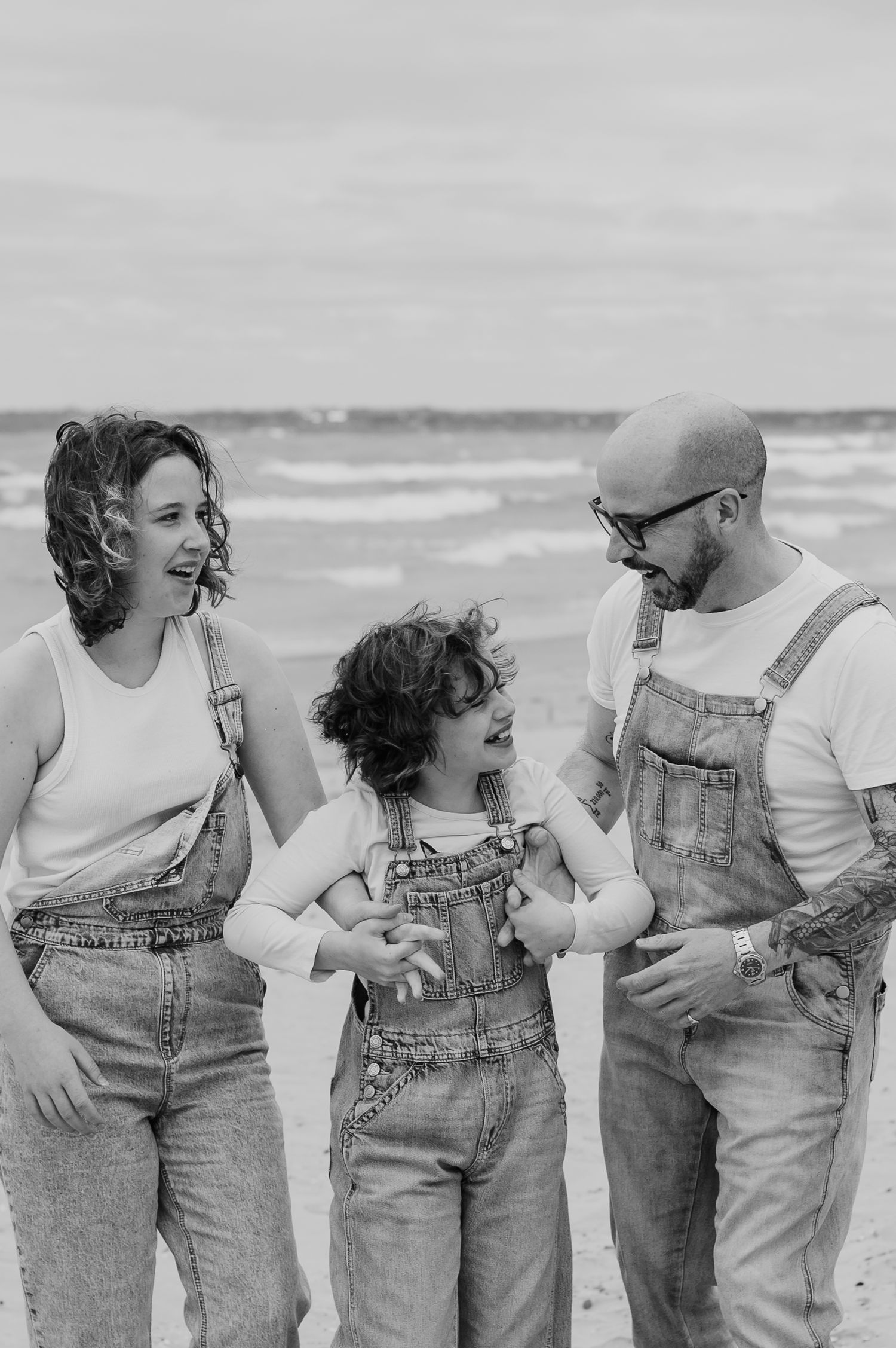 A family wearing matching denim overalls shares a laugh together on a cloudy beach.