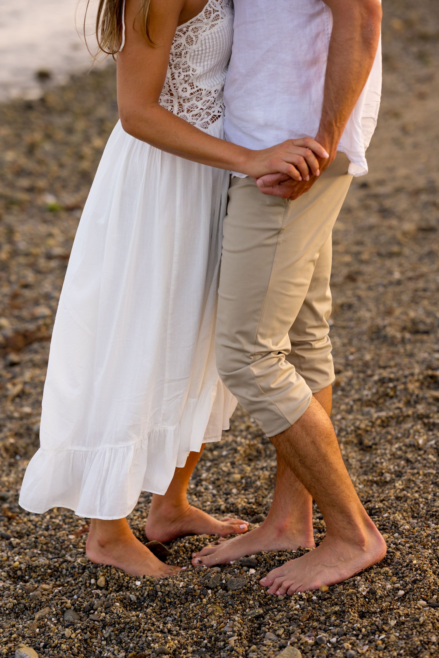 Barefoot couple stands close together on sandy ground with her wearing a white dress and him in khaki shorts.