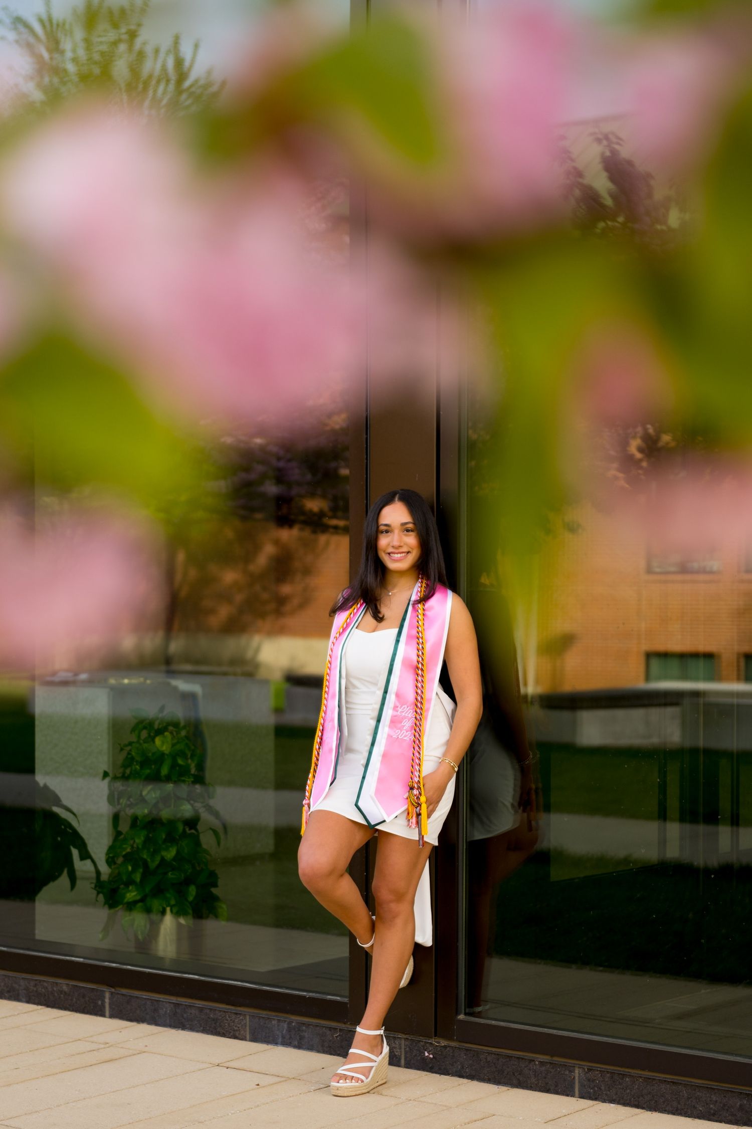 Sacred Heart University graduate poses by reflective building with pink cherry blossoms in foreground.