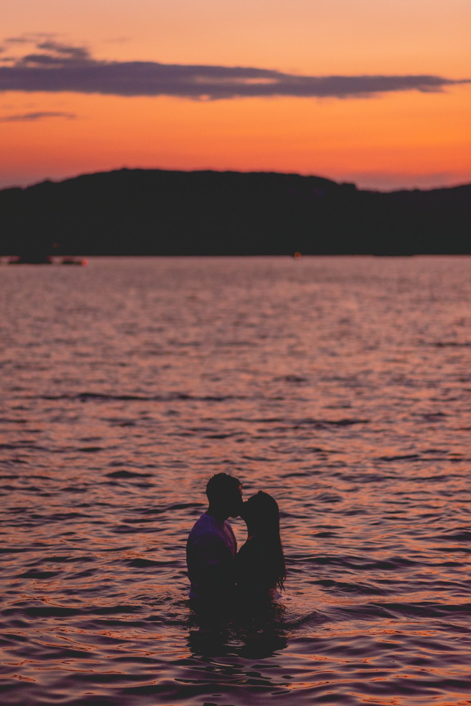 Silhouettes of couple floating in water against vibrant orange sunset.