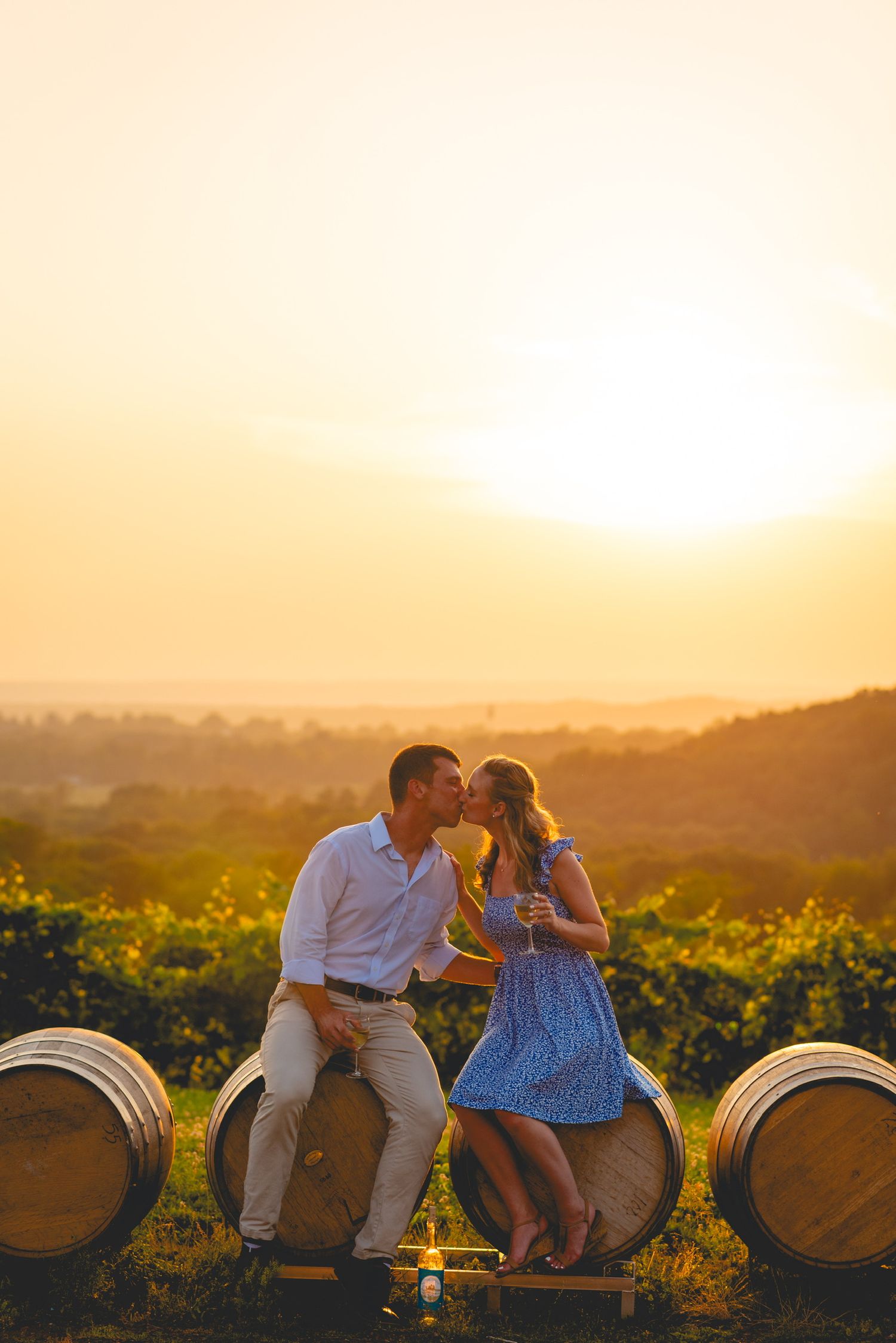 Couple sitting on wine barrels in vineyard during golden sunset.