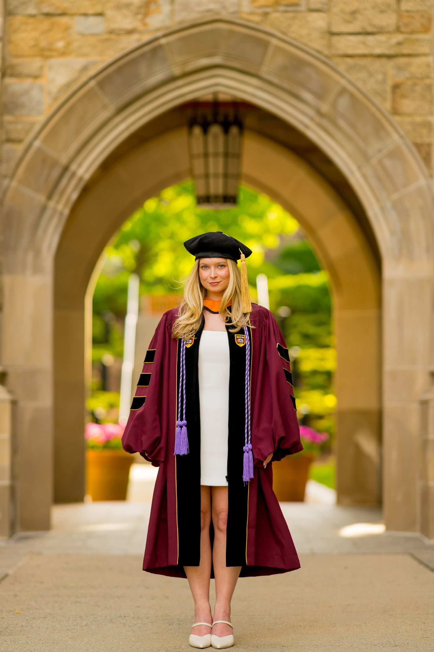 Graduate in burgundy academic regalia poses under stone archways.