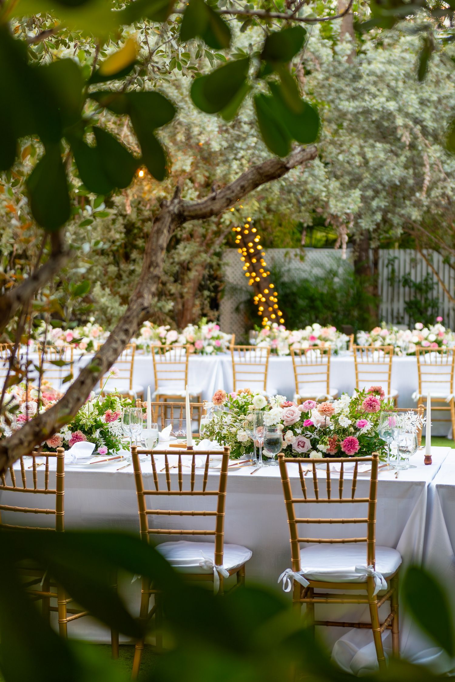 Outdoor wedding reception setup with white tables, wooden chairs, and string lights under blooming trees in Miami, Florida.