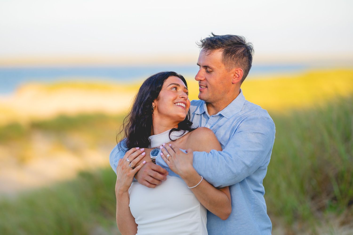 Romantic couple embraces in golden sunlit field during sunset photoshoot at Chatham Lighthouse Beach in Chatham, Massachusetts.