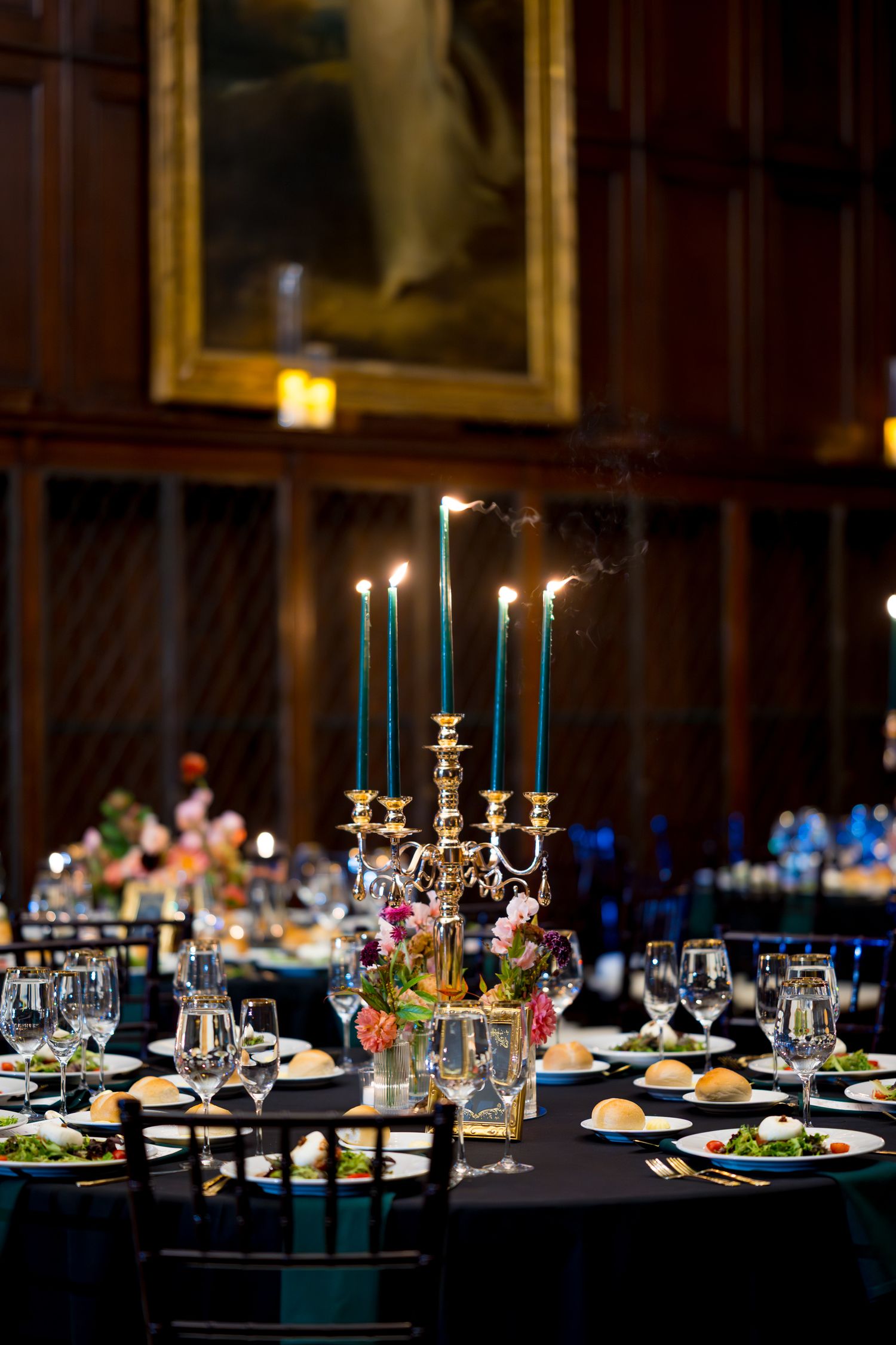 Elegant wedding reception tables set with tall blue candles, fine china, and floral centerpieces in a dimly lit venue.