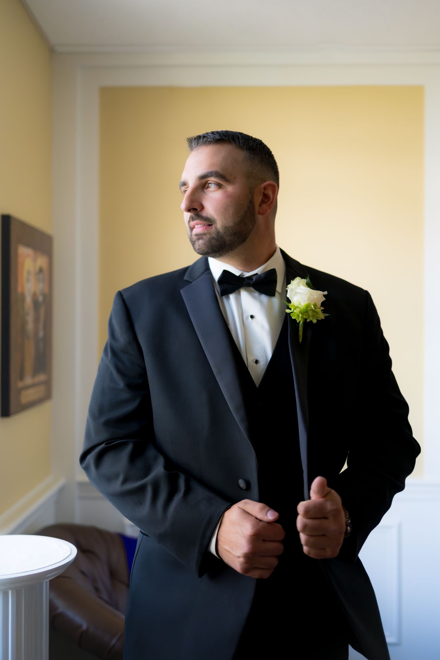 A groom in a classic black tuxedo adjusts his bowtie while getting ready for his wedding ceremony.