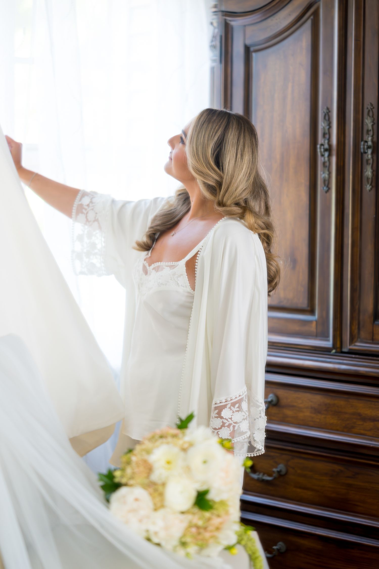 A bride in a white dress holds a bouquet while standing in front of a wooden armoire.