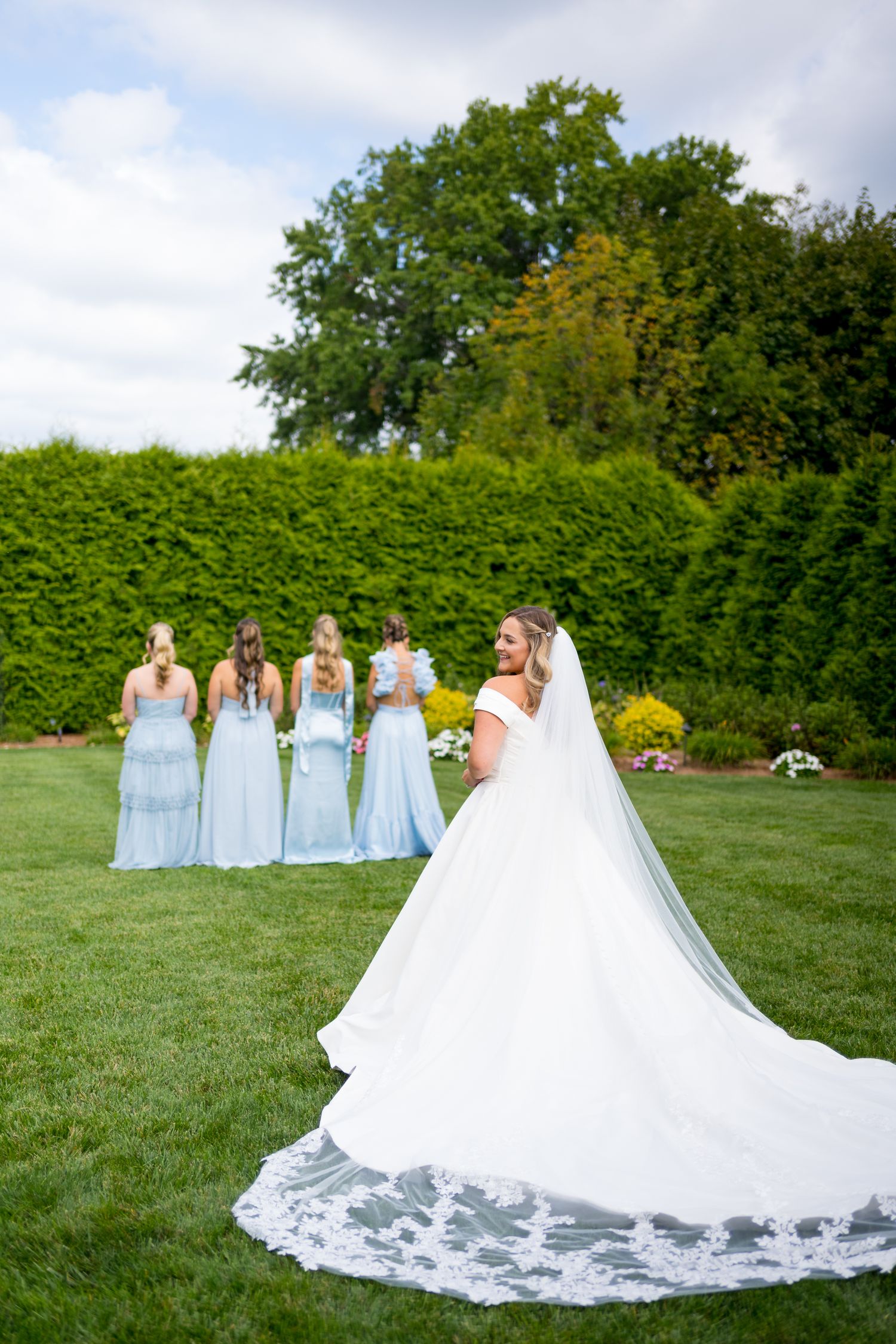 A bridal party in light blue dresses poses on a green lawn with flowing white wedding gown train.