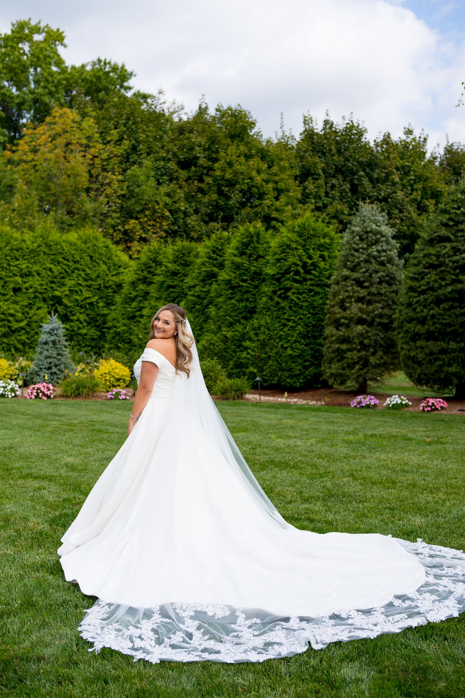 A stunning white wedding dress with cathedral train spreads across green lawn against garden backdrop.
