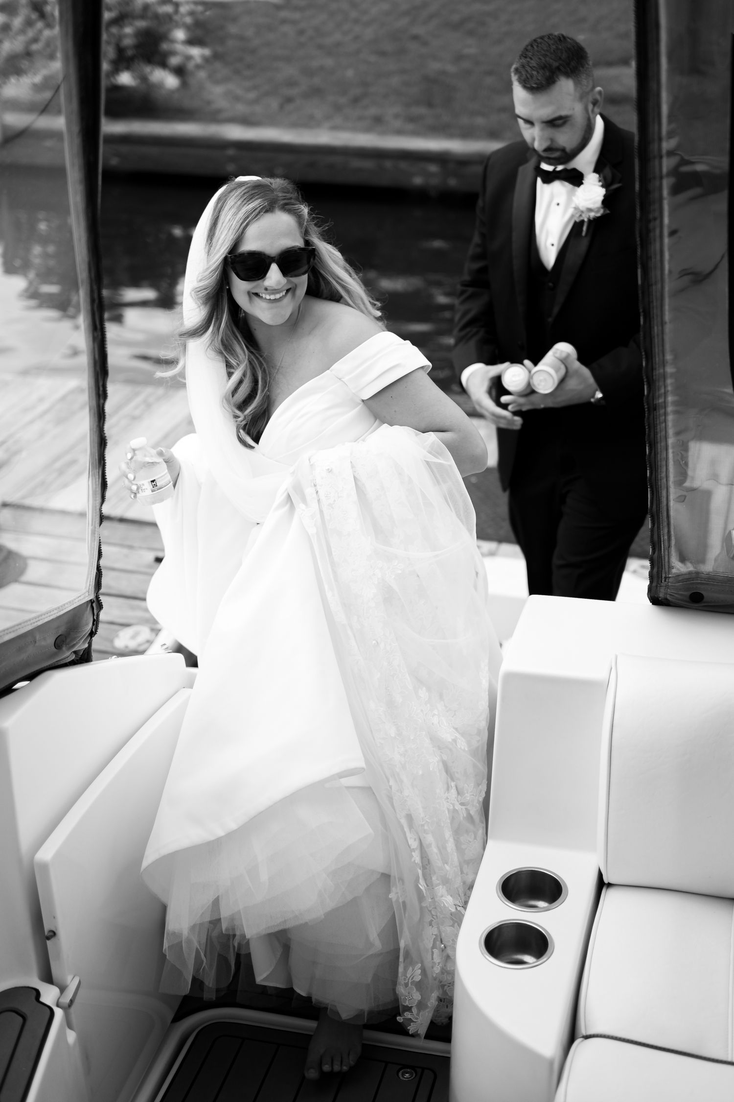 Bride in sunglasses and off-shoulder wedding dress stepping onto a boat while groom watches in black tuxedo.