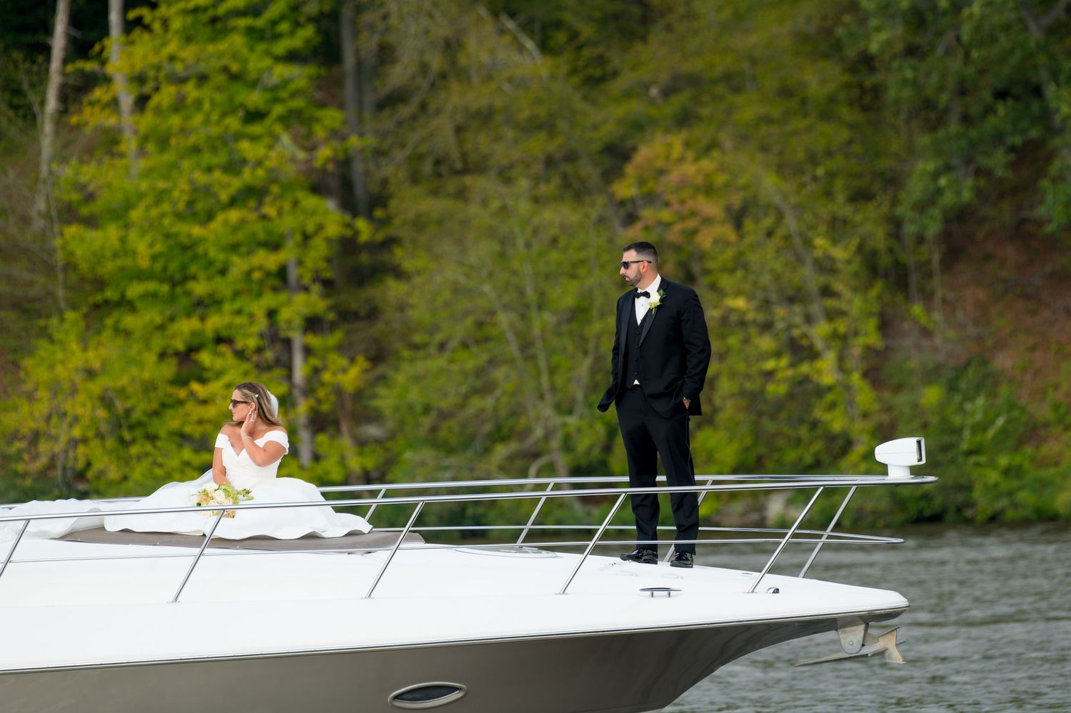 A couple in formal wedding attire stands on the bow of a luxury yacht with lush green forest in the background.