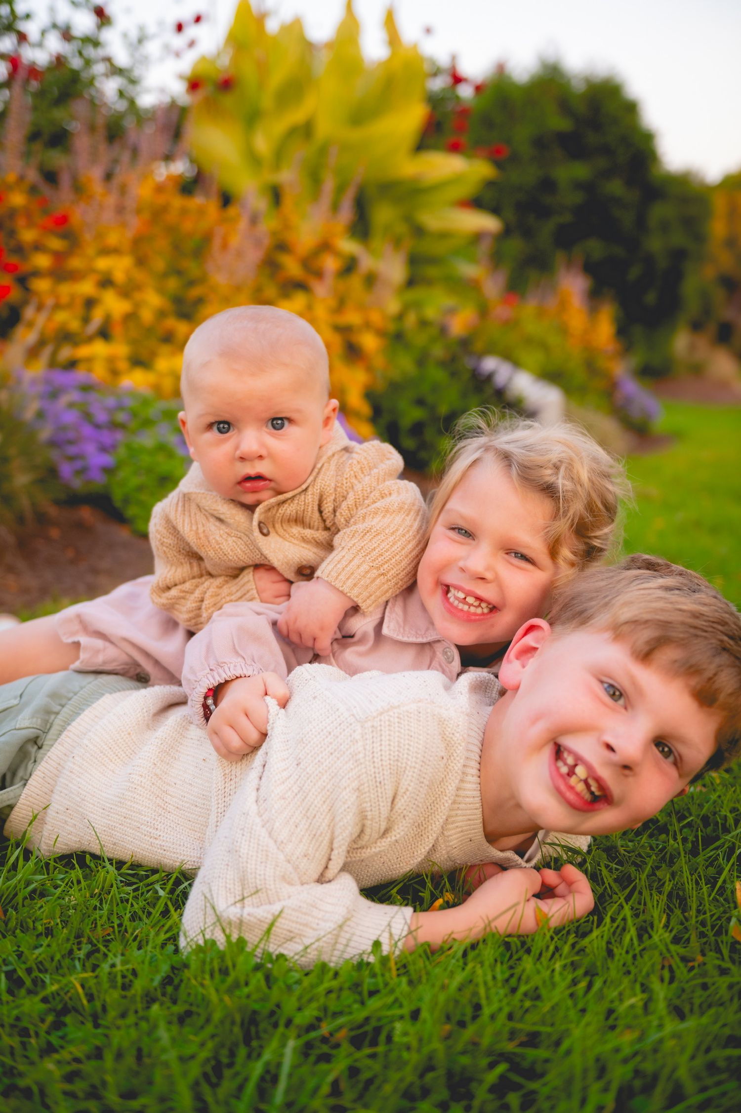 Two young children in cream sweaters playfully lie on green grass during an autumn day.