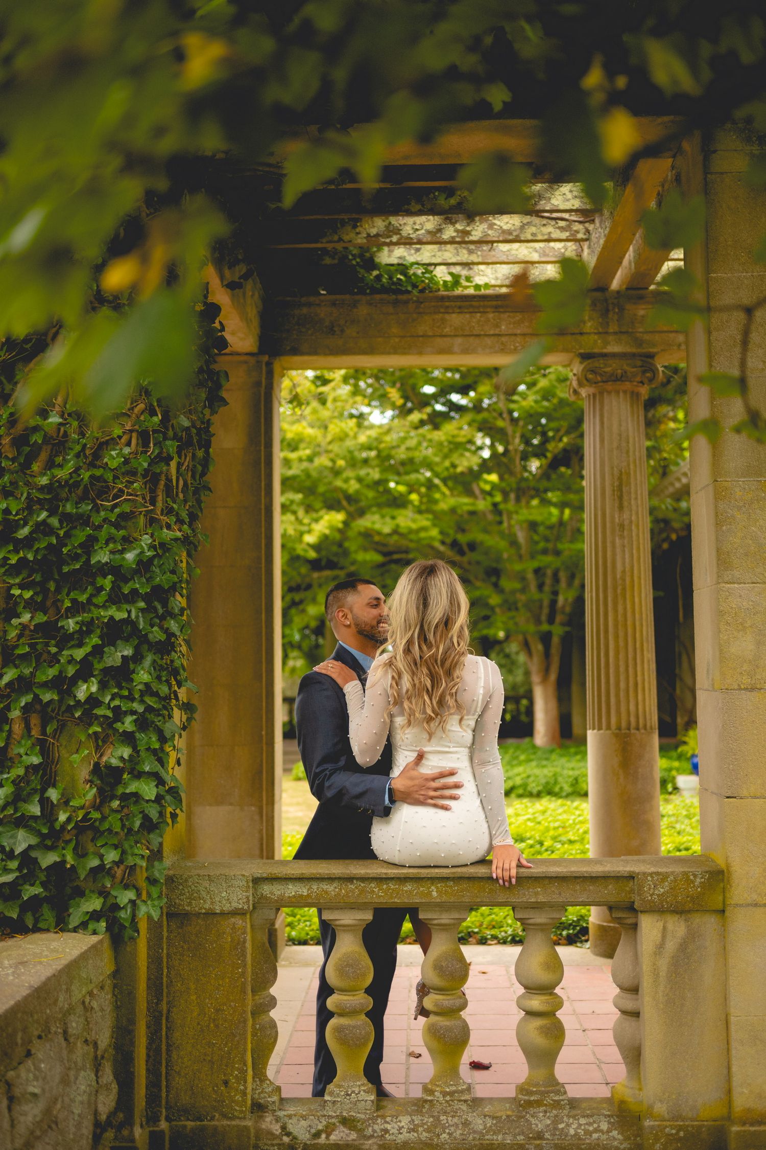 A couple shares an intimate moment in an ornate garden pergola surrounded by greenery.
