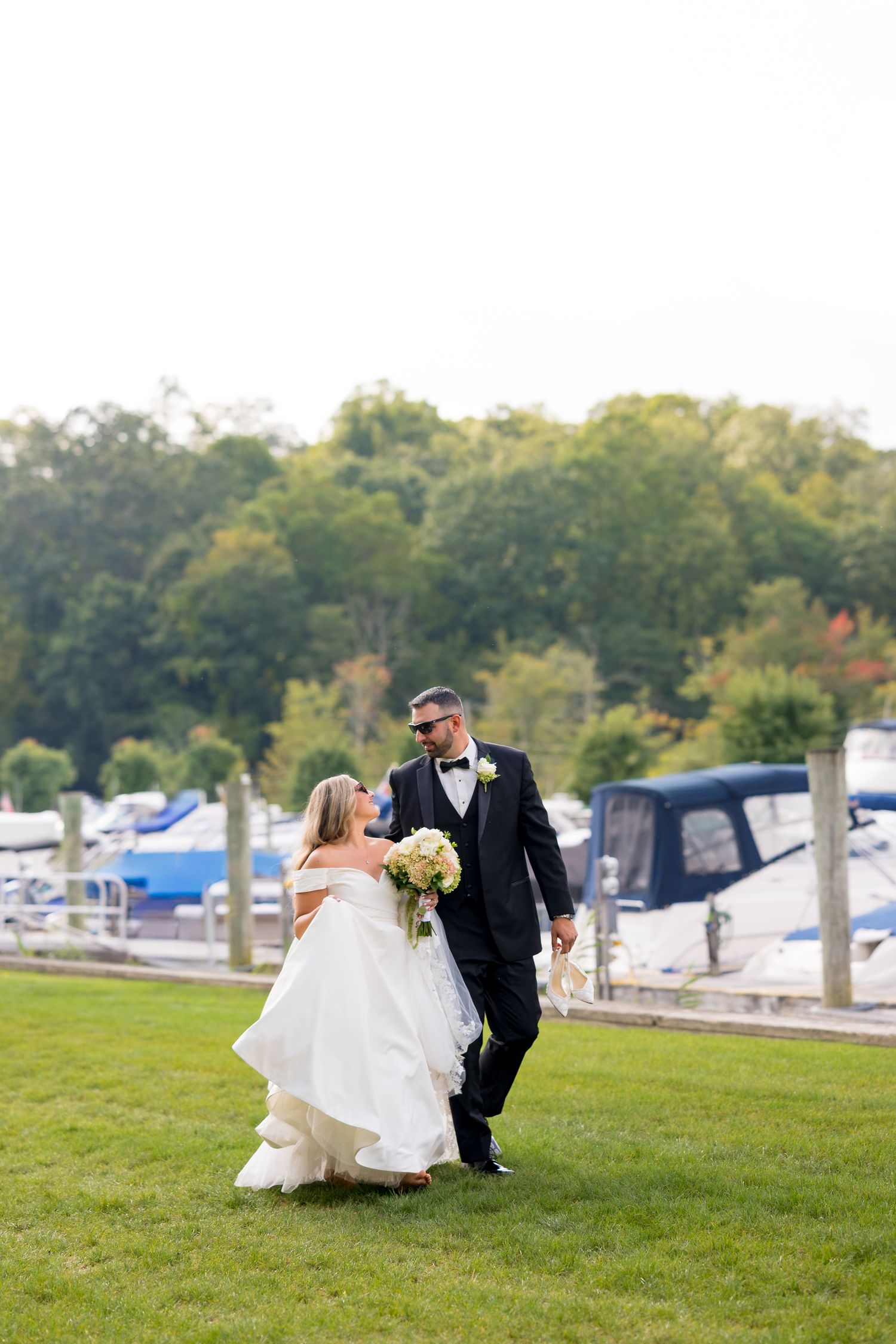 Newlyweds playfully posing on grass near a marina with boats in background.