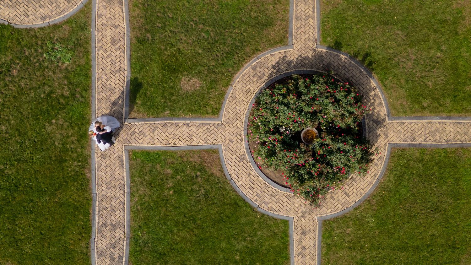 Aerial view of a car driving past a circular garden with stone pathways and green grass in a geometric pattern.