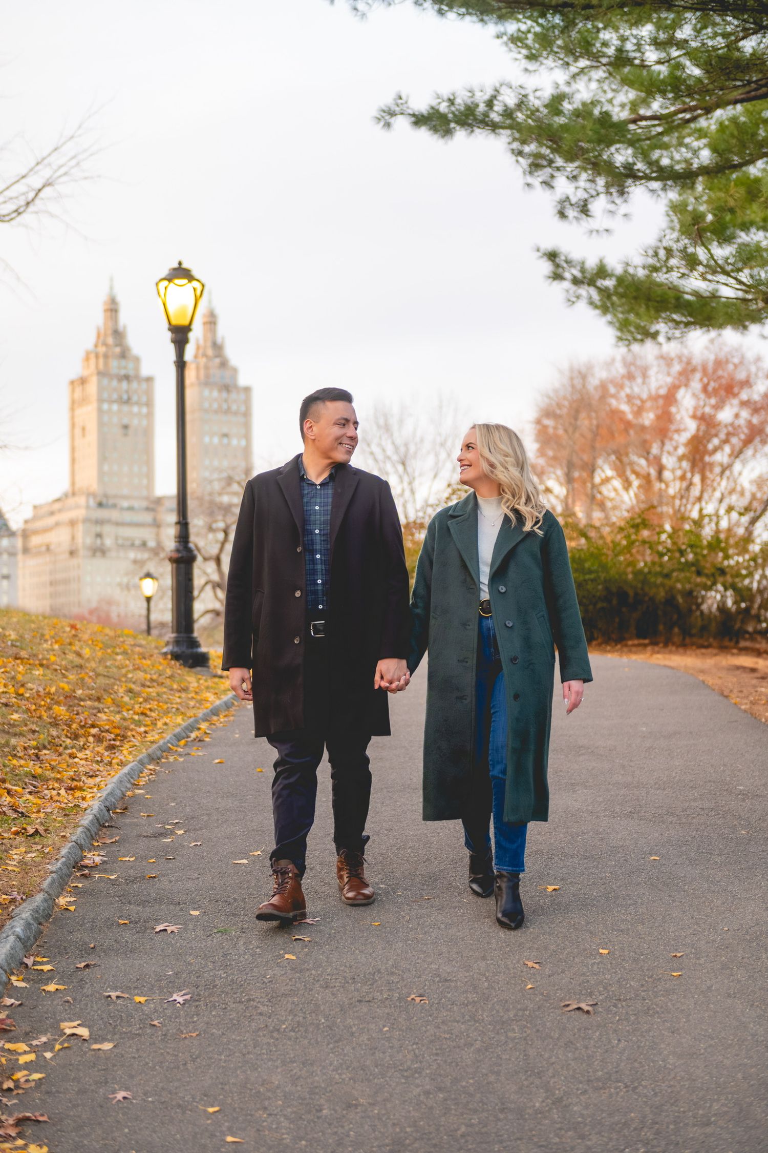 A couple takes an autumn stroll along a path with city buildings in the background in Central Park.