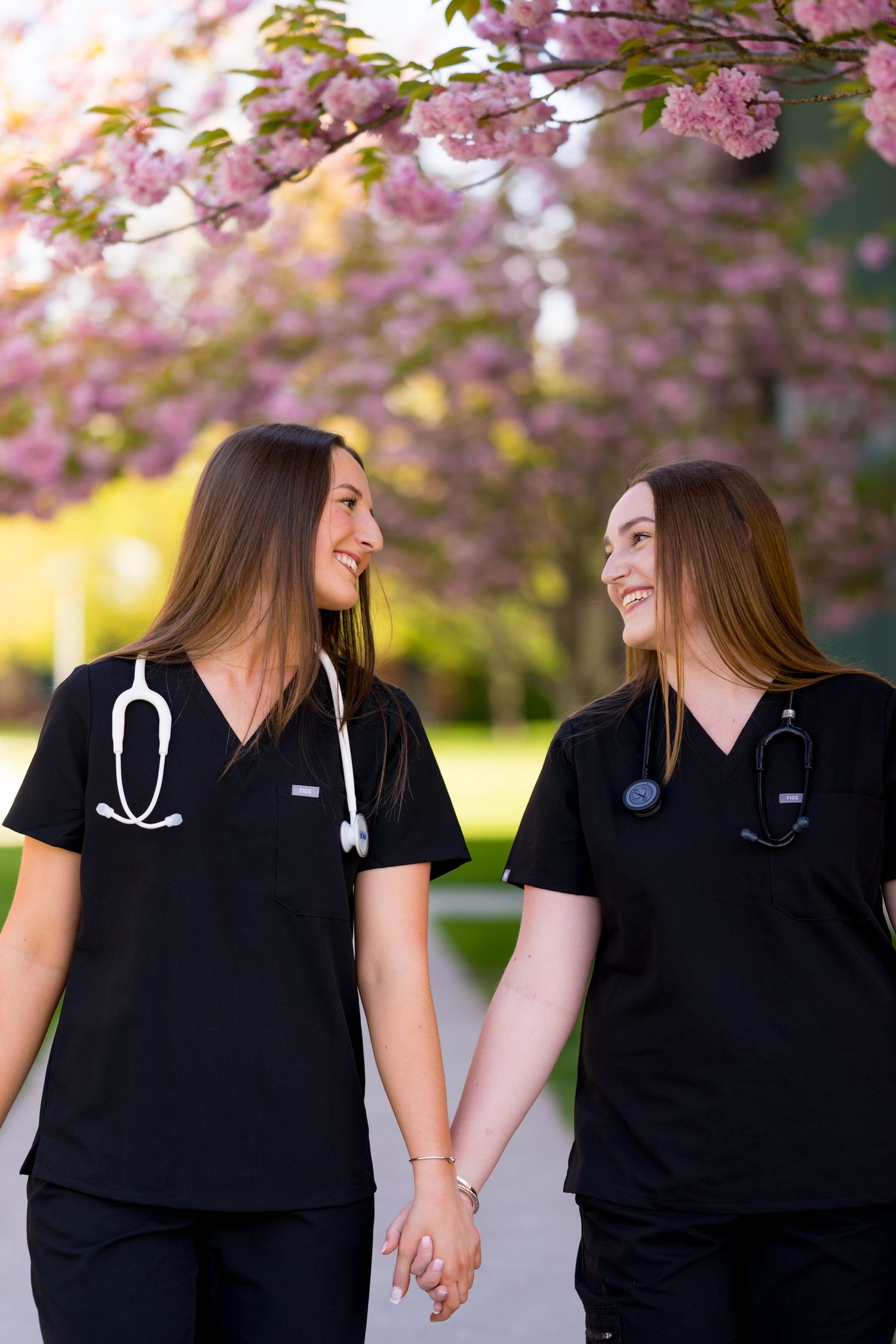 Two Sacred Heart University nurses in black scrubs walking together under blooming cherry blossom trees.