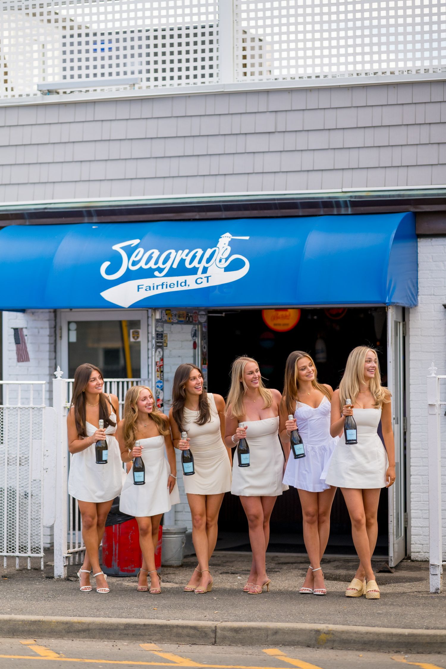 Fairfield University graduates standing together in white dresses posing against Seagrape Cafe for graduation photos.