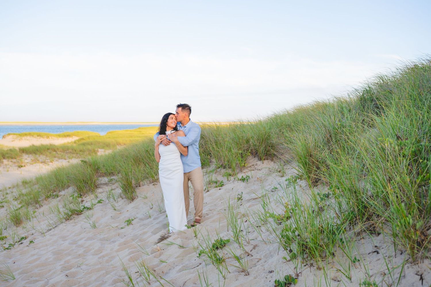 Romantic couple embracing on Chatham Lighthouse Beach at sunset with tall grass and ocean in background.