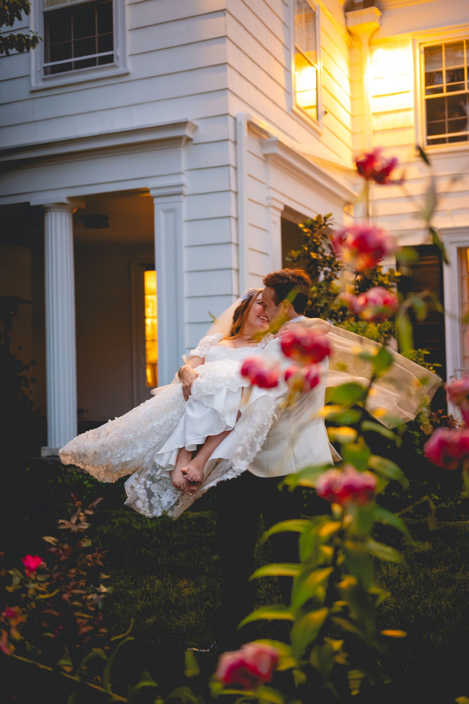 White wedding dress twirling in motion against a warmly lit house exterior with pink flowers.