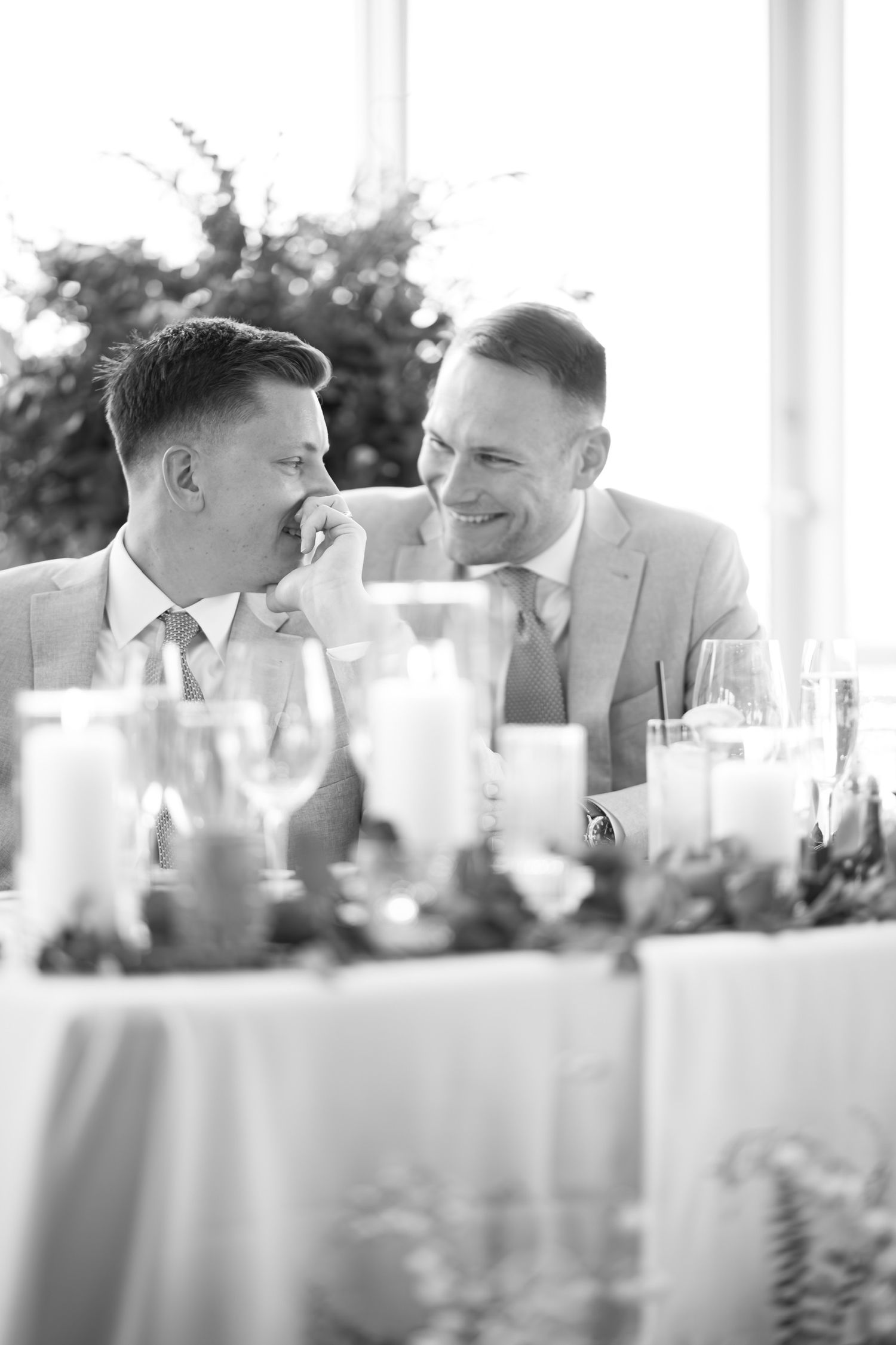 Two men in formal attire share a candid laughing moment at a reception table.