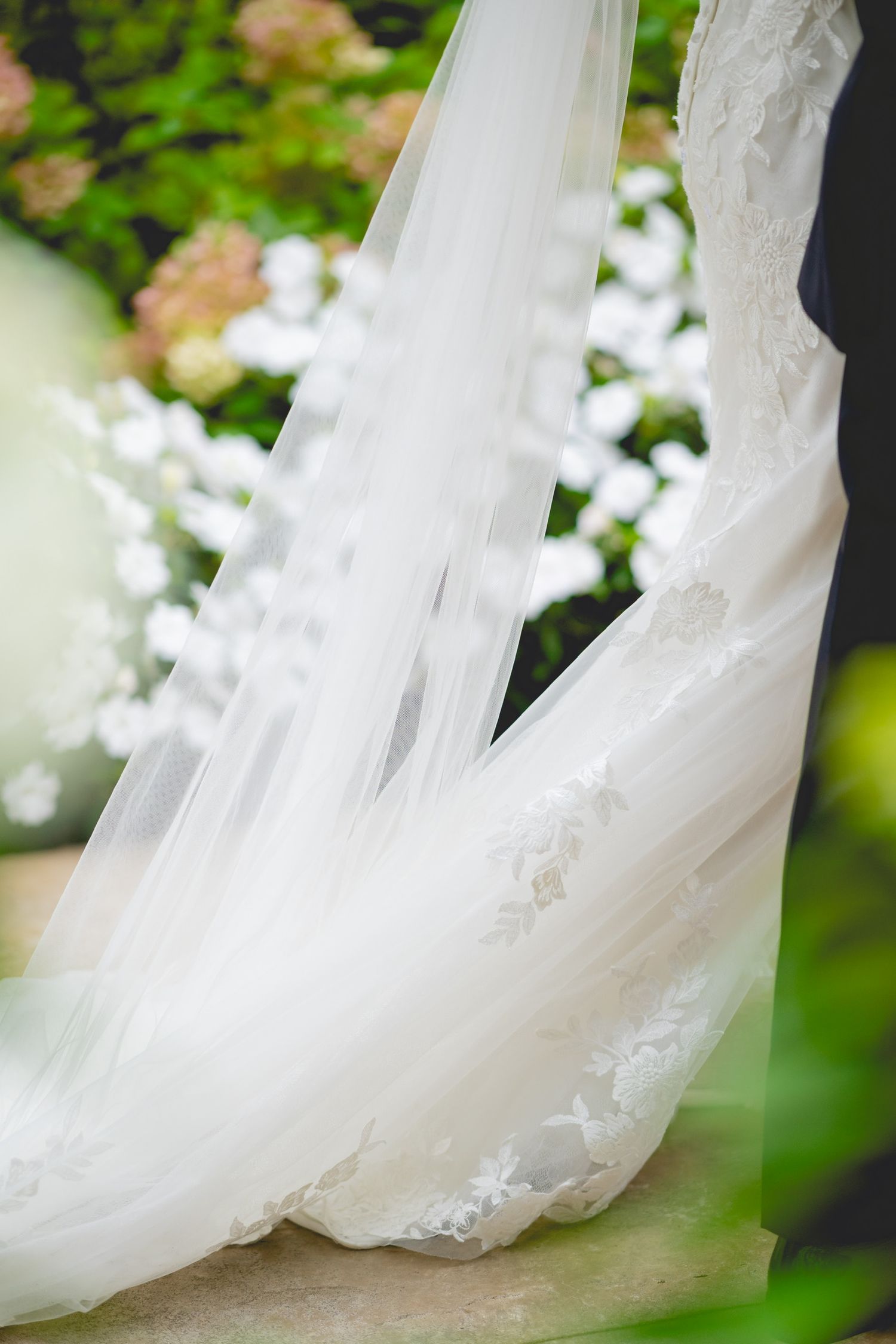 Close-up detail of flowing white wedding dress veil with delicate lace trim against green foliage.