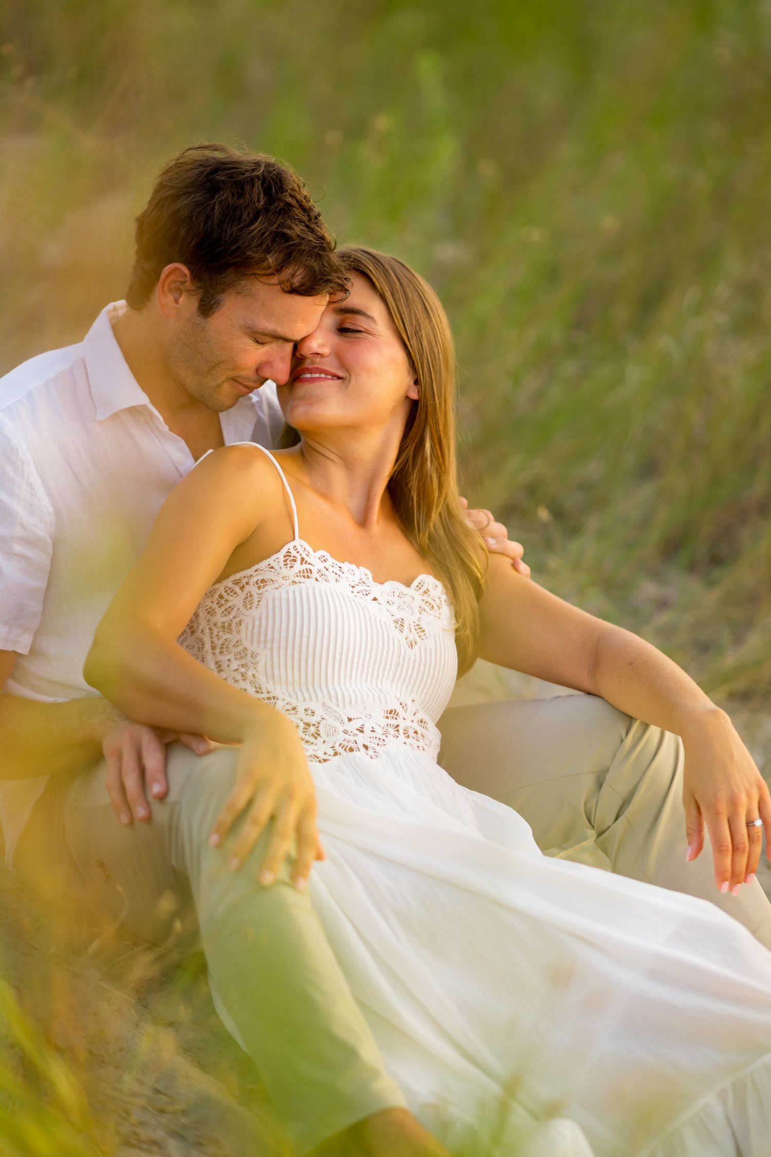 Romantic couple embraces on beach in white summer outfits during golden hour photoshoot.
