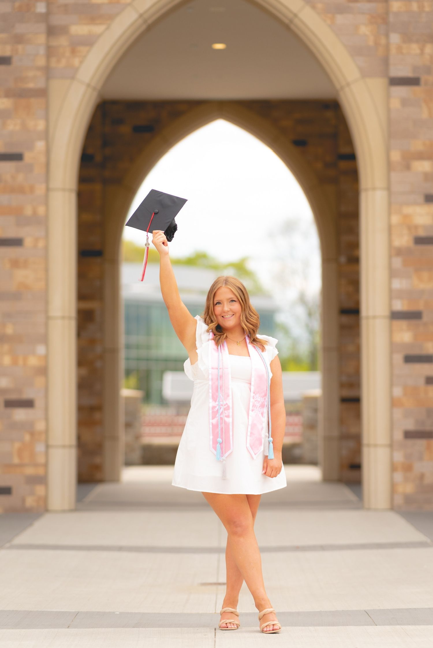 Figure in white dress and graduation cap stands under stone archways.