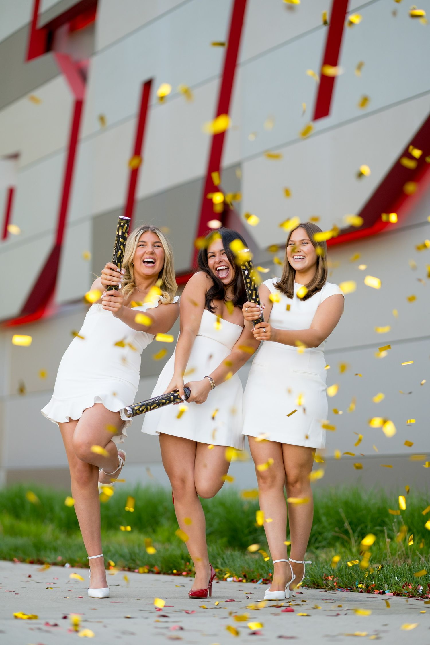 Sacred Heart University graduates in white dresses celebrate with confetti and champagne outdoors.