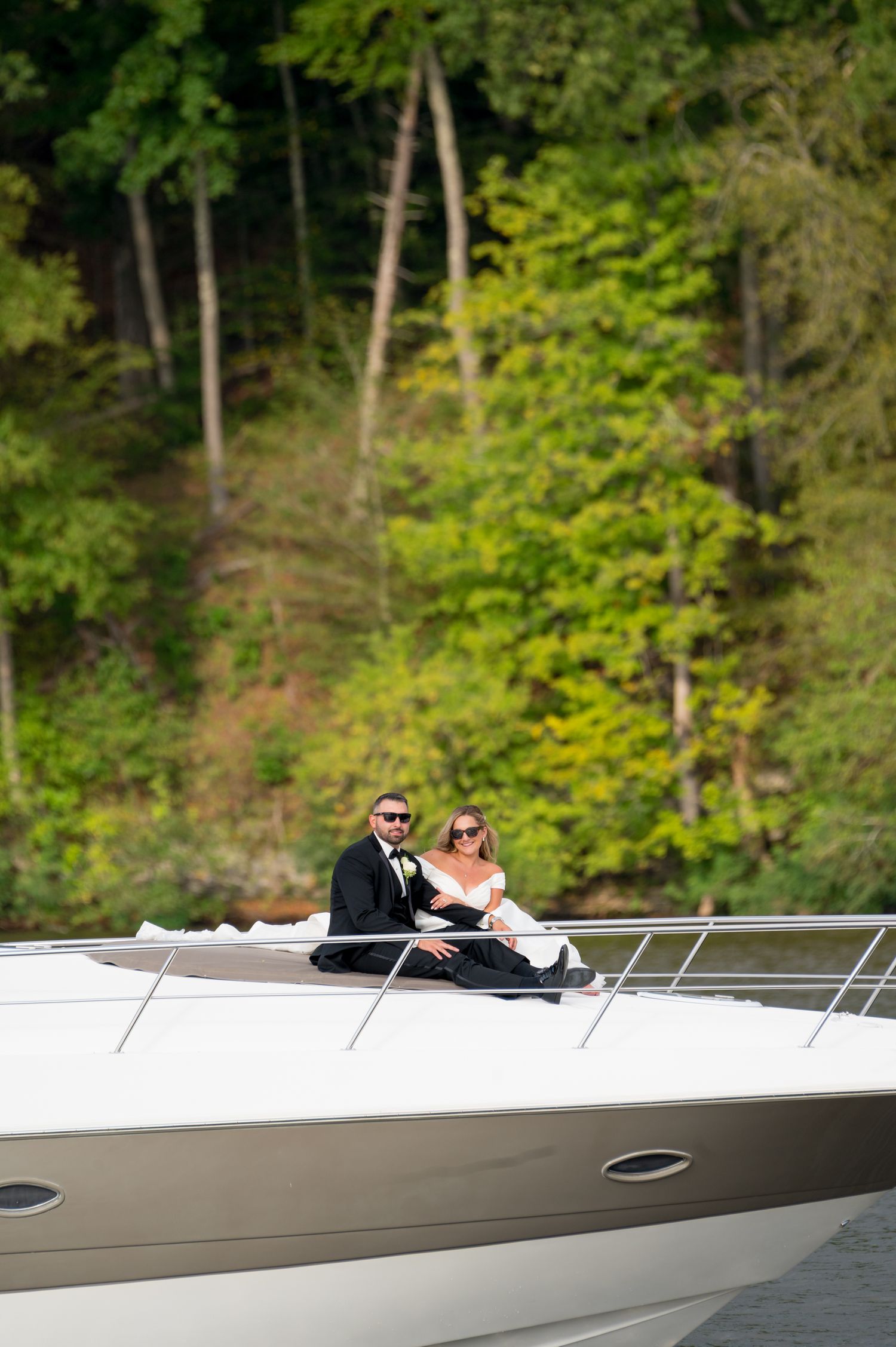 A couple relaxes on the deck of a luxury yacht surrounded by lush green trees along the shoreline.