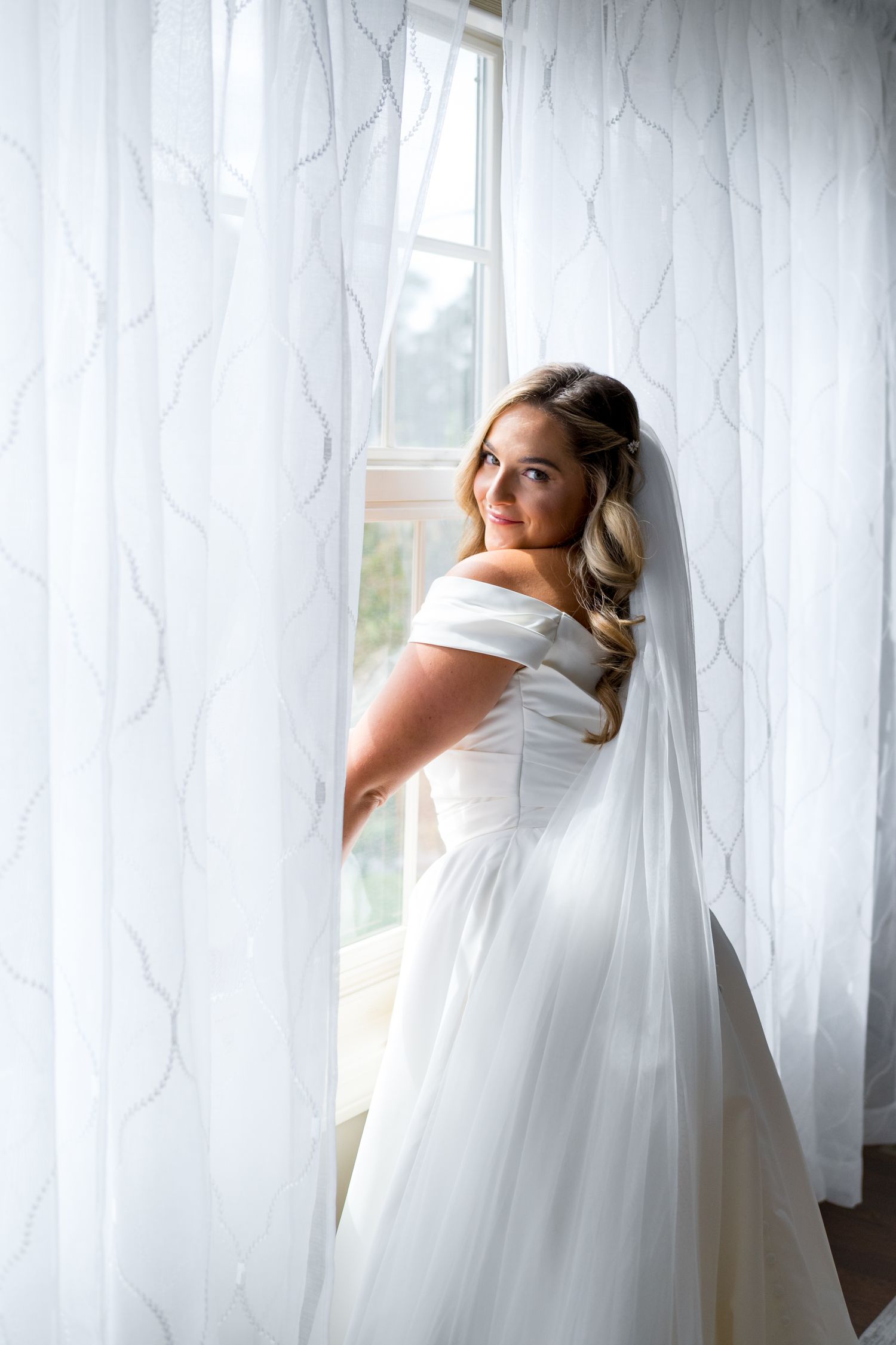 Bride in flowing white wedding dress and veil poses elegantly against sheer white curtains in natural window light.