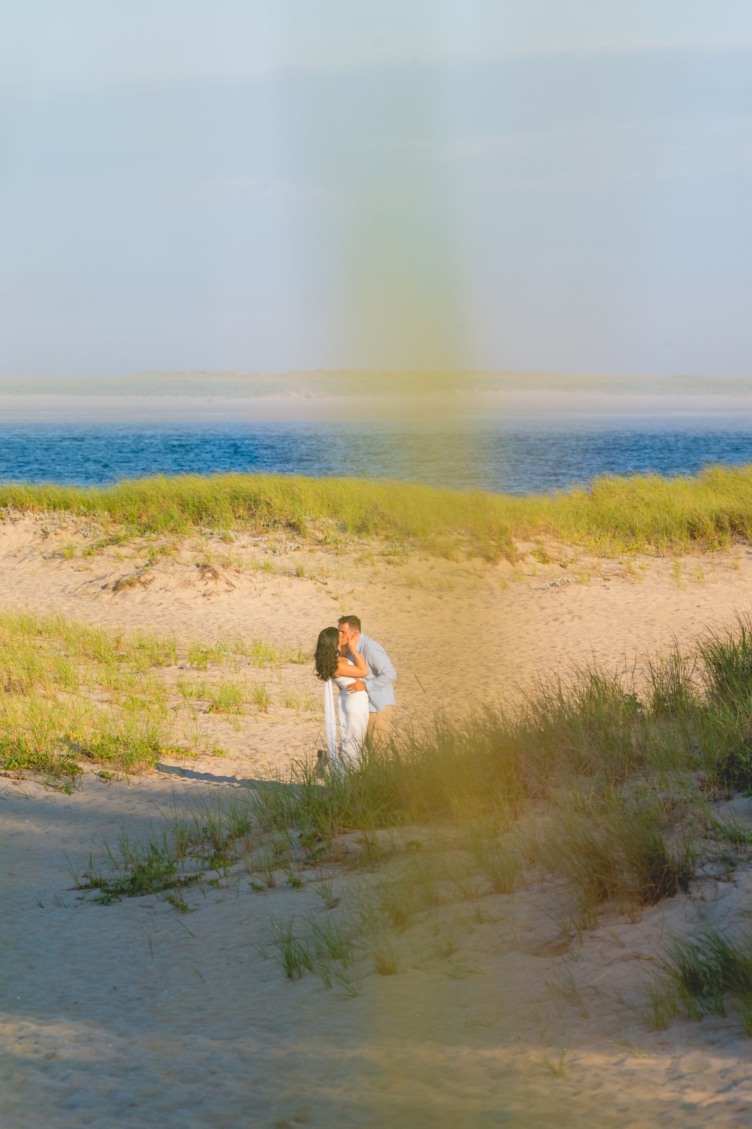 Romantic couple embraces on a sandy beach with tall grass and ocean views at sunset.
