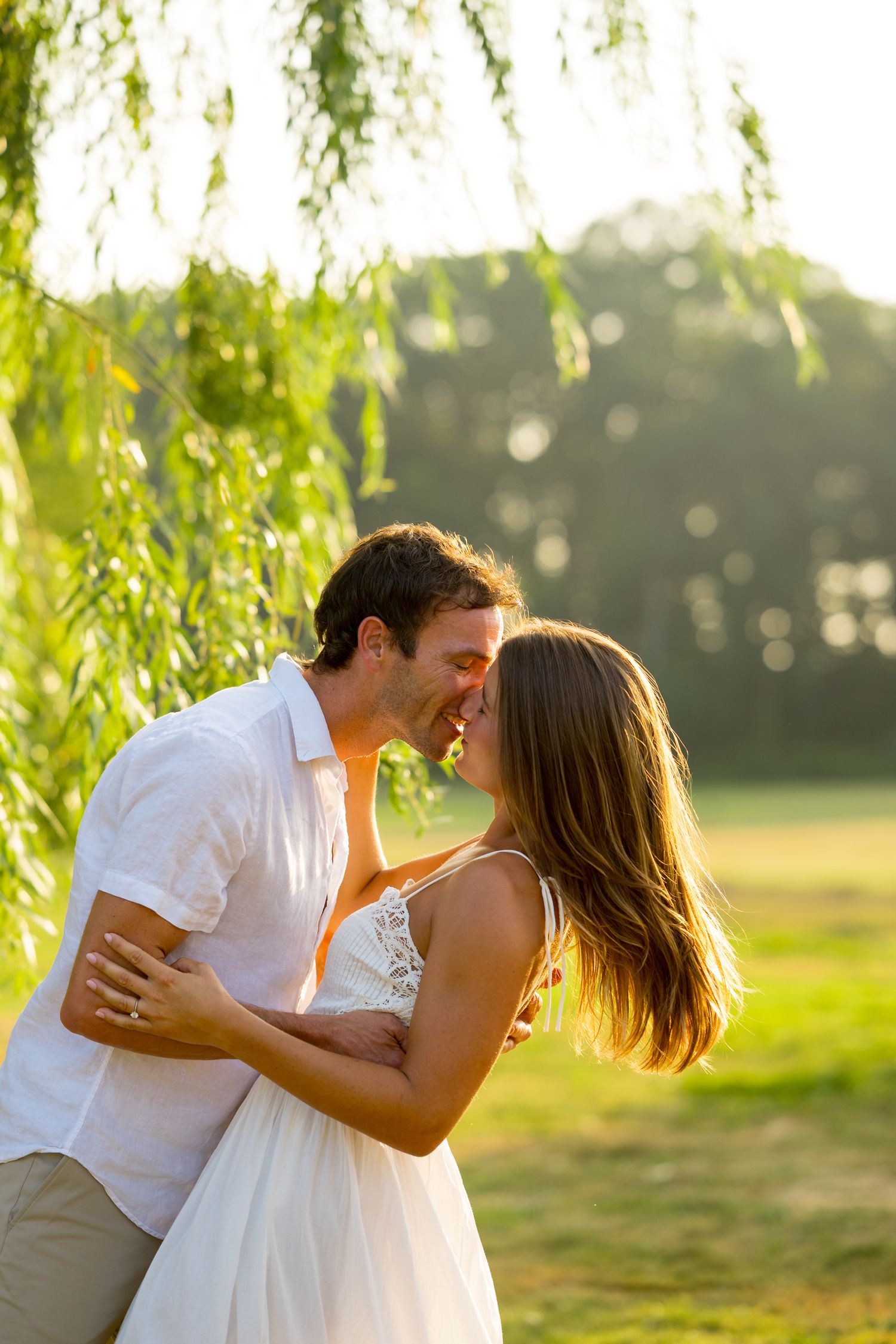 A romantic couple shares intimate moments under a weeping willow tree during golden hour sunset.