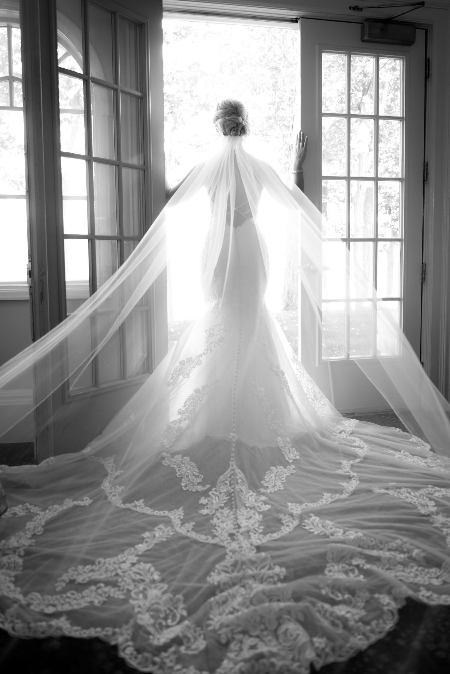 Elegant white wedding dress with intricate lace train displayed in window-lit room.