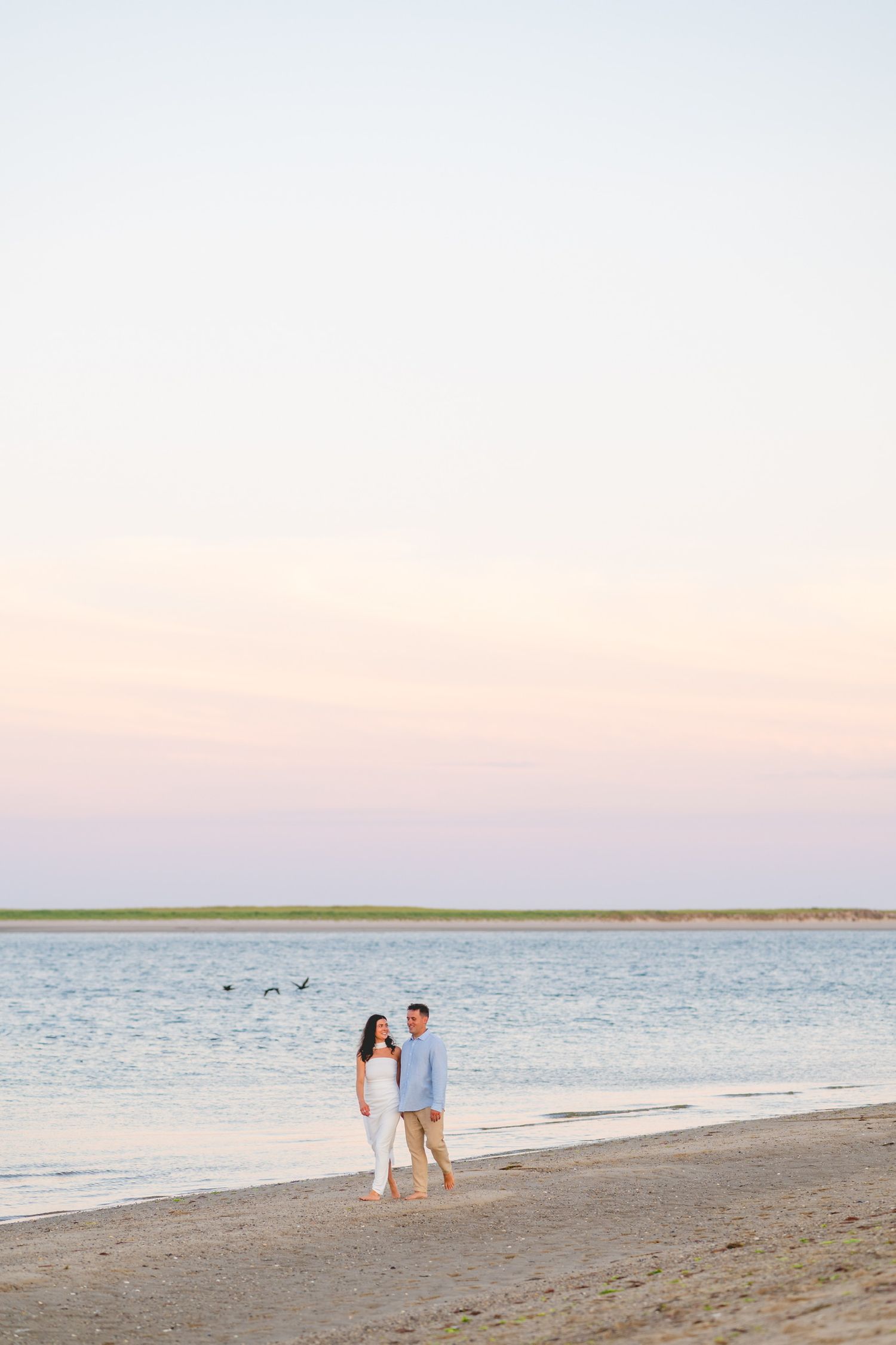A couple walks along a serene Cape Cod beach at sunset with calm waters and pink sky in the background.